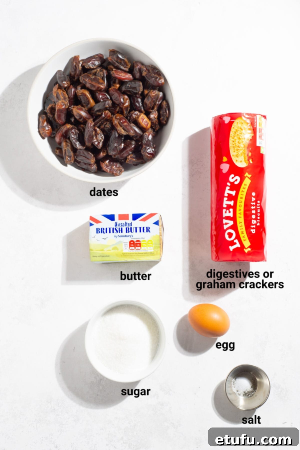 Effortless Date Bars 3 A collection of ingredients for date squares, including butter, dates, sugar, egg, and digestive biscuits, neatly arranged on a white surface.