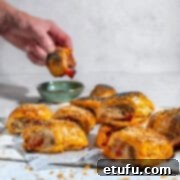 Mini sausage rolls on a white background with a hand dipping a sausage roll into tomato sauce.