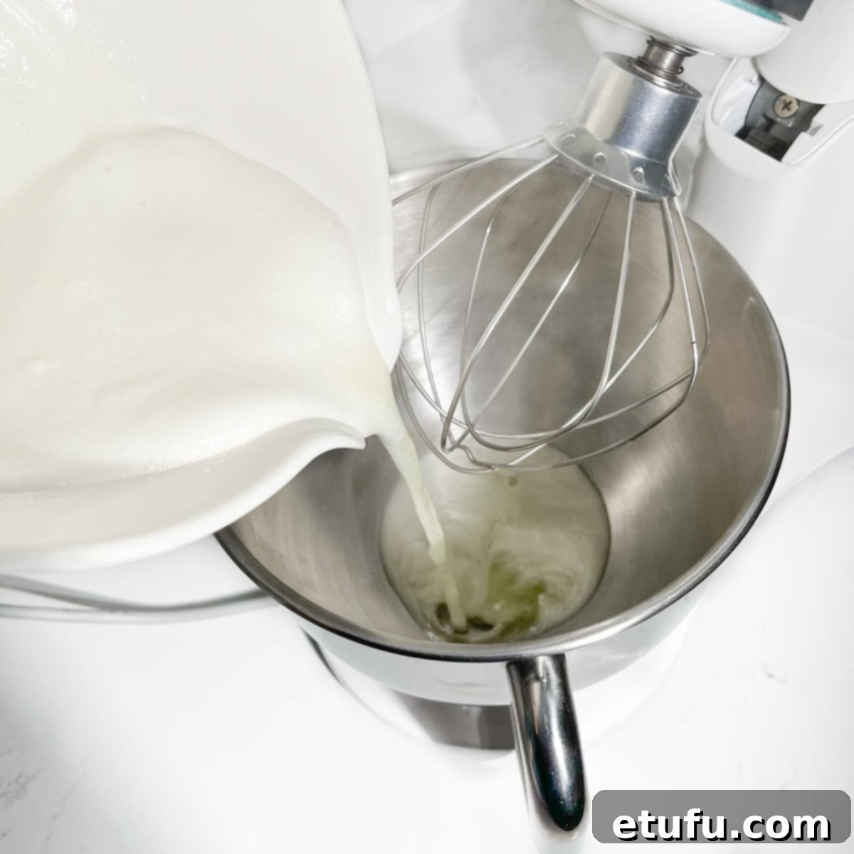 The hot egg and sugar mixture being carefully poured into the clean, grease-free bowl of a stand mixer.