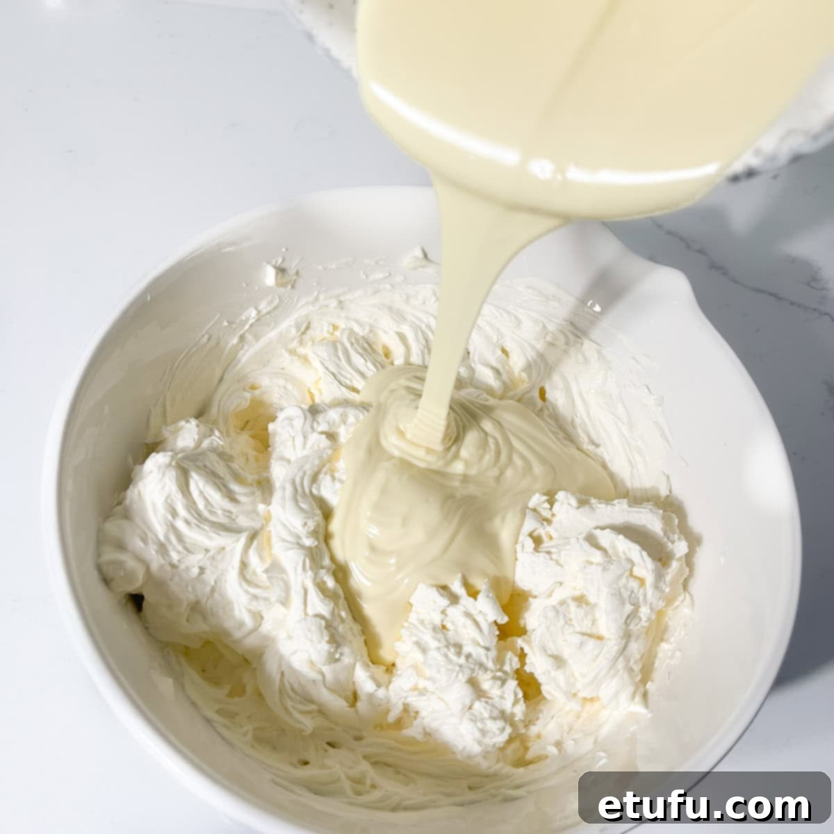 Pouring the slightly cooled melted white chocolate into the combined cream, cream cheese, and mascarpone mixture.