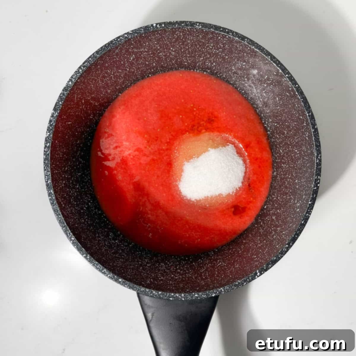 Adding granulated sugar to the strawberry puree in a small black saucepan over low heat.