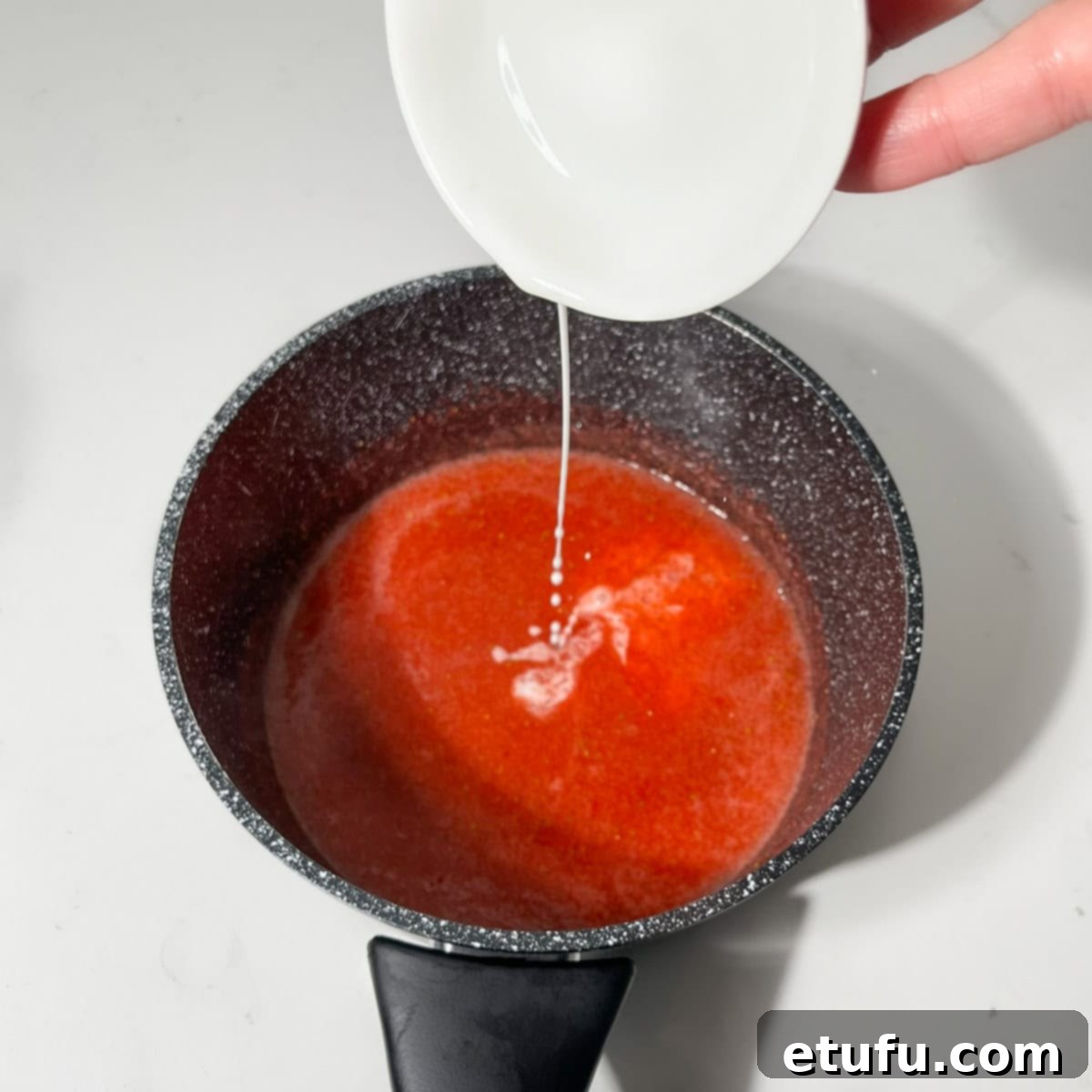 Whisking the cornstarch slurry into the simmering strawberry sauce in a small black saucepan.