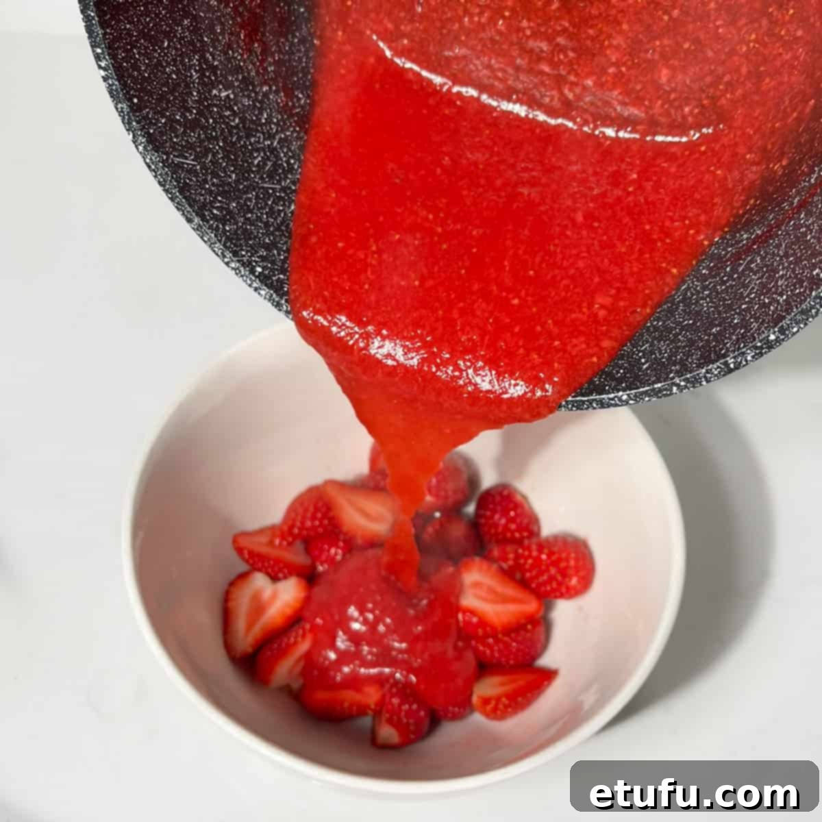 Pouring the hot, thickened strawberry sauce over the fresh, sliced strawberries in a white bowl.