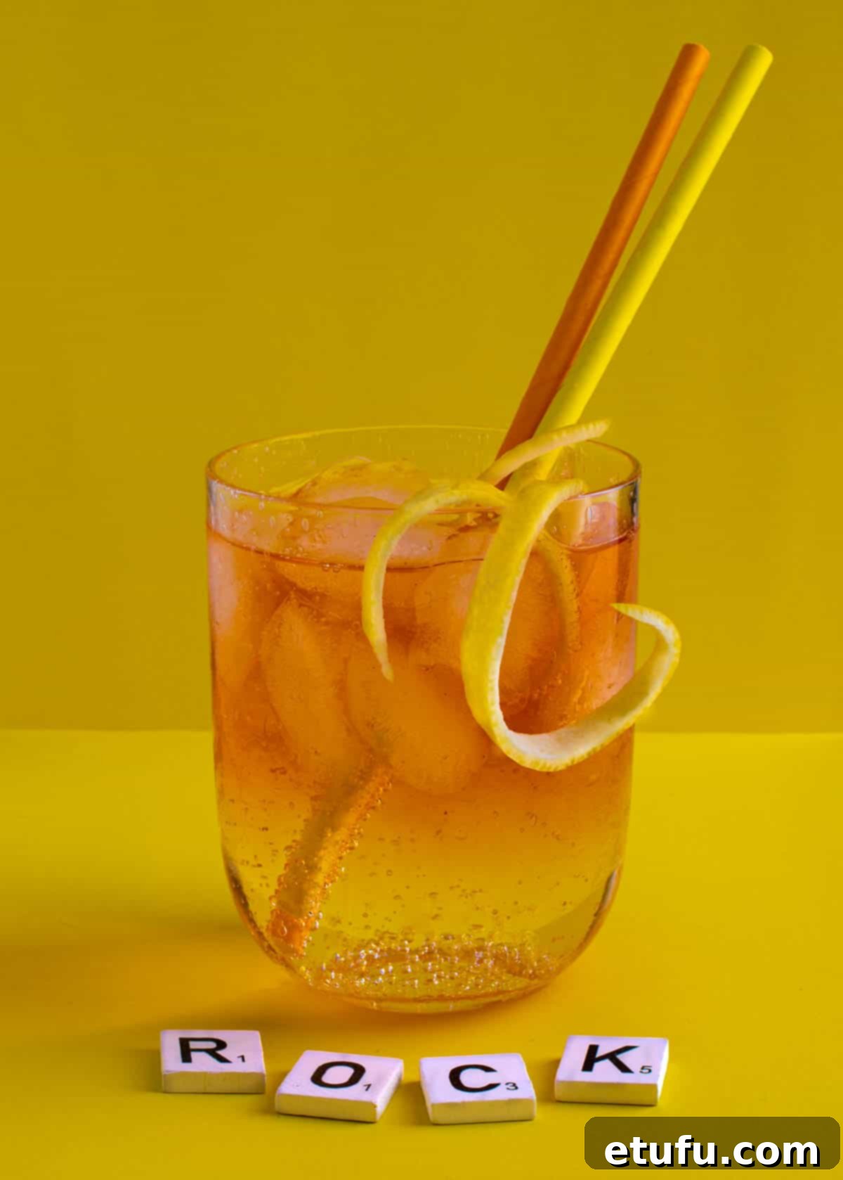 A medium glass of Rock shandy on a yellow background with an orange and yellow straw, highlighting its vibrant colors and refreshing appeal.