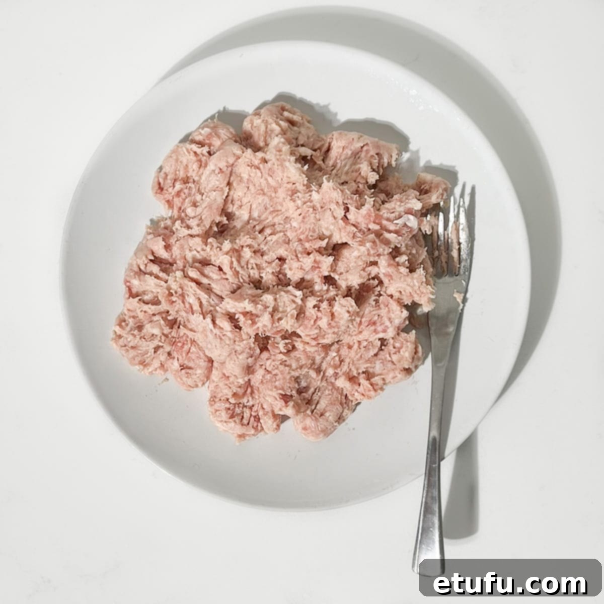 Raw sausage meat being broken apart with a fork on a white plate, ready to be incorporated into the savory filling.