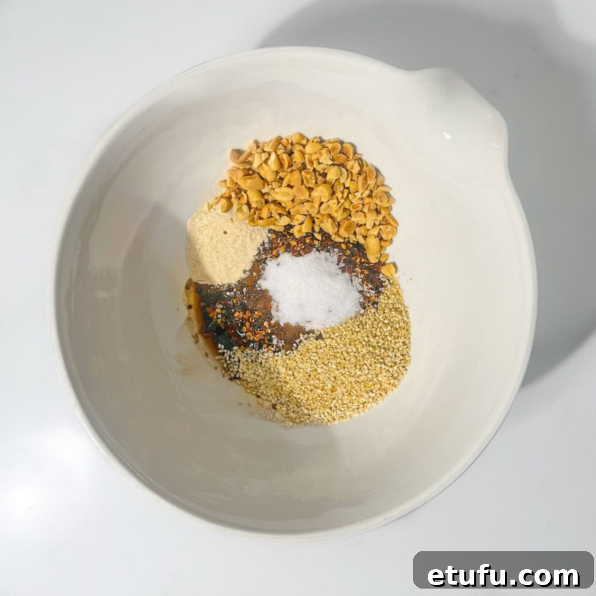 A large white bowl containing chilli flakes, salt, sugar, mushroom powder, soy sauce, rice vinegar, sesame seeds, and chopped roasted peanuts, ready to be mixed.