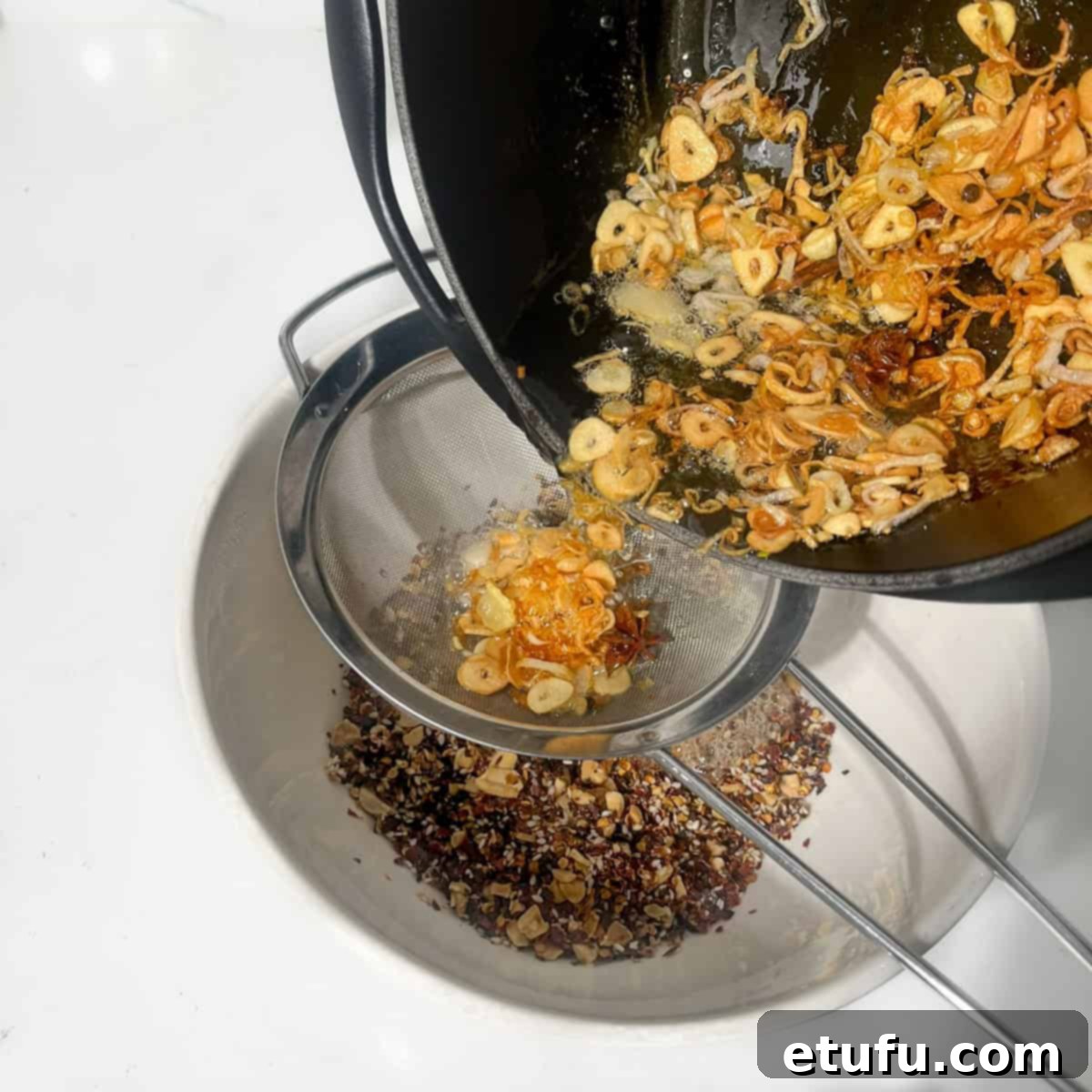 Hot infused oil is being carefully poured through a sieve, directly onto the prepared dry chilli base in the large heatproof bowl. Steam rises as the oil sizzles.