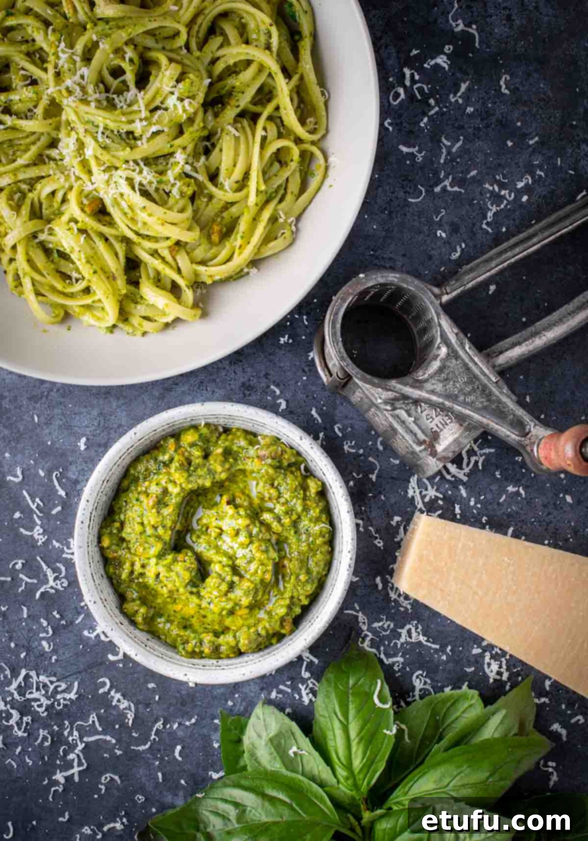 A bowl of pistachio pesto surrounded by a bowl of pasta, a parmesan grater and parmesan cheese.