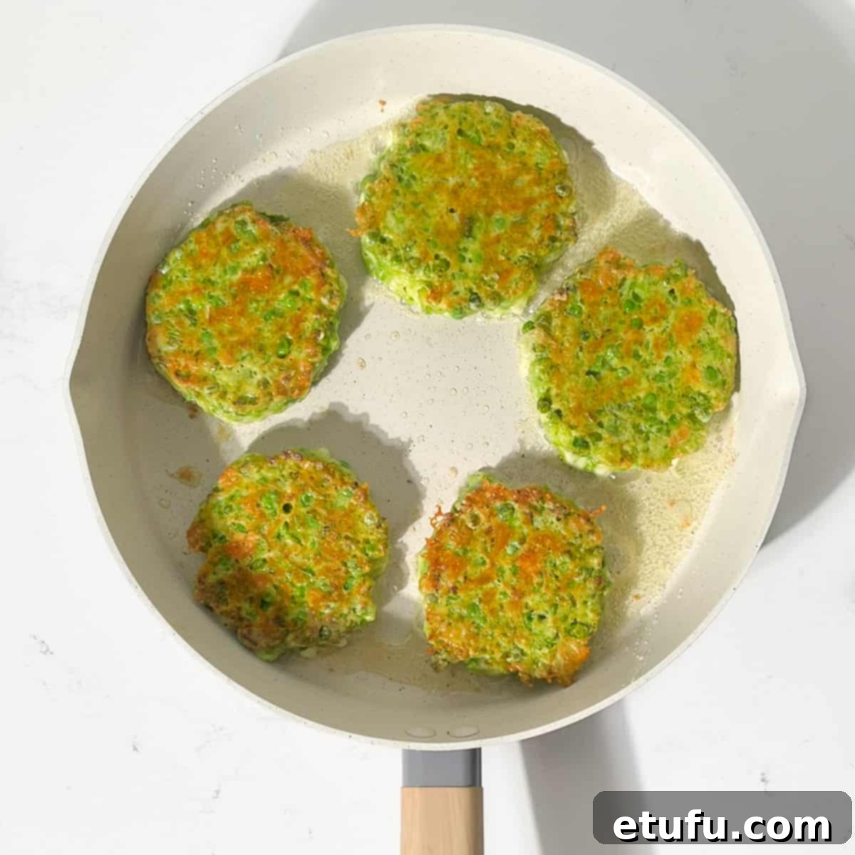 Several green pea fritters sizzling in a large frying pan, showing a golden-brown and crispy exterior.