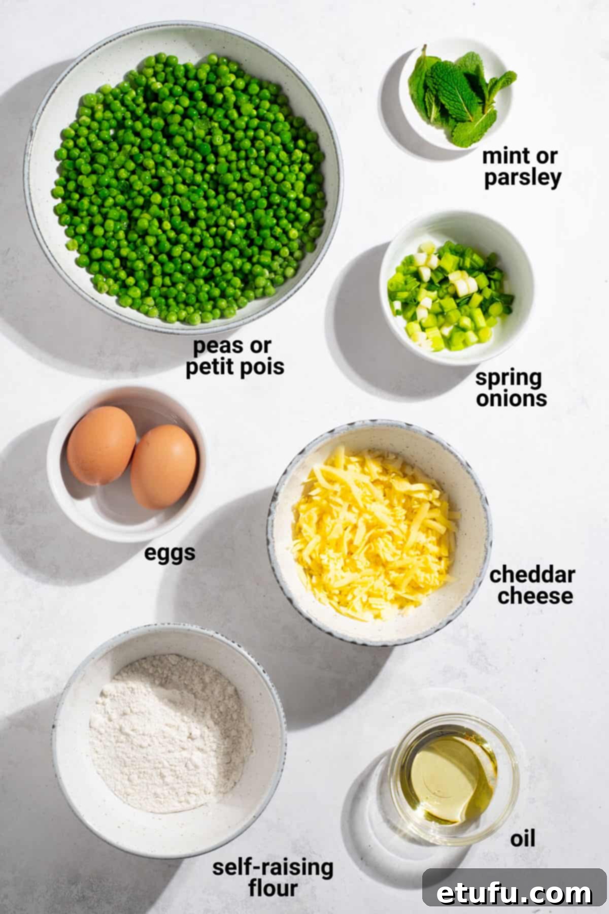 A selection of fresh ingredients laid out on a wooden board, including peas, spring onions, cheese, flour, eggs, and herbs, ready for making green pea fritters.