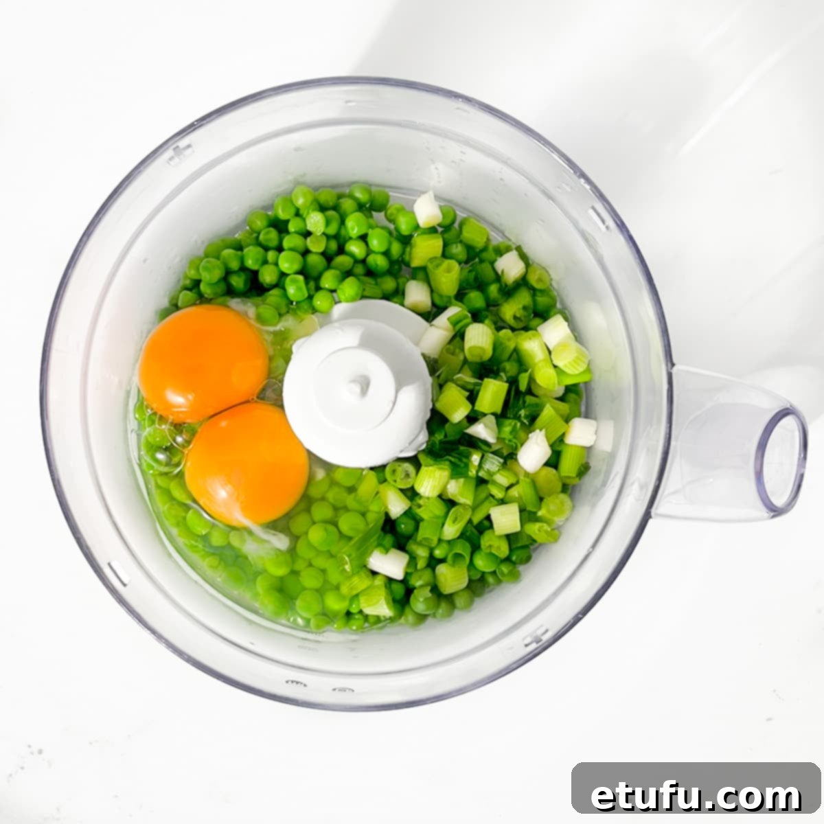 Half of the cooked peas, two large eggs, and chopped spring onions being added to the bowl of a food processor for blending.