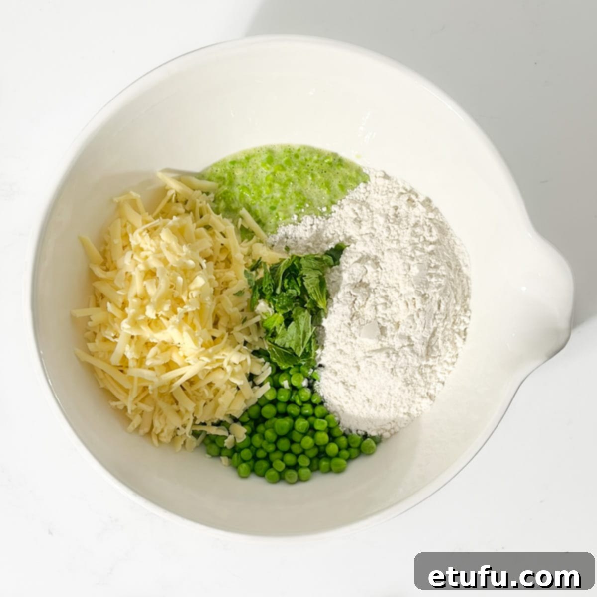 All the remaining ingredients, including the other half of the peas, grated cheese, self-raising flour, chopped mint, and salt, being added to the blended pea mixture in a large white bowl.