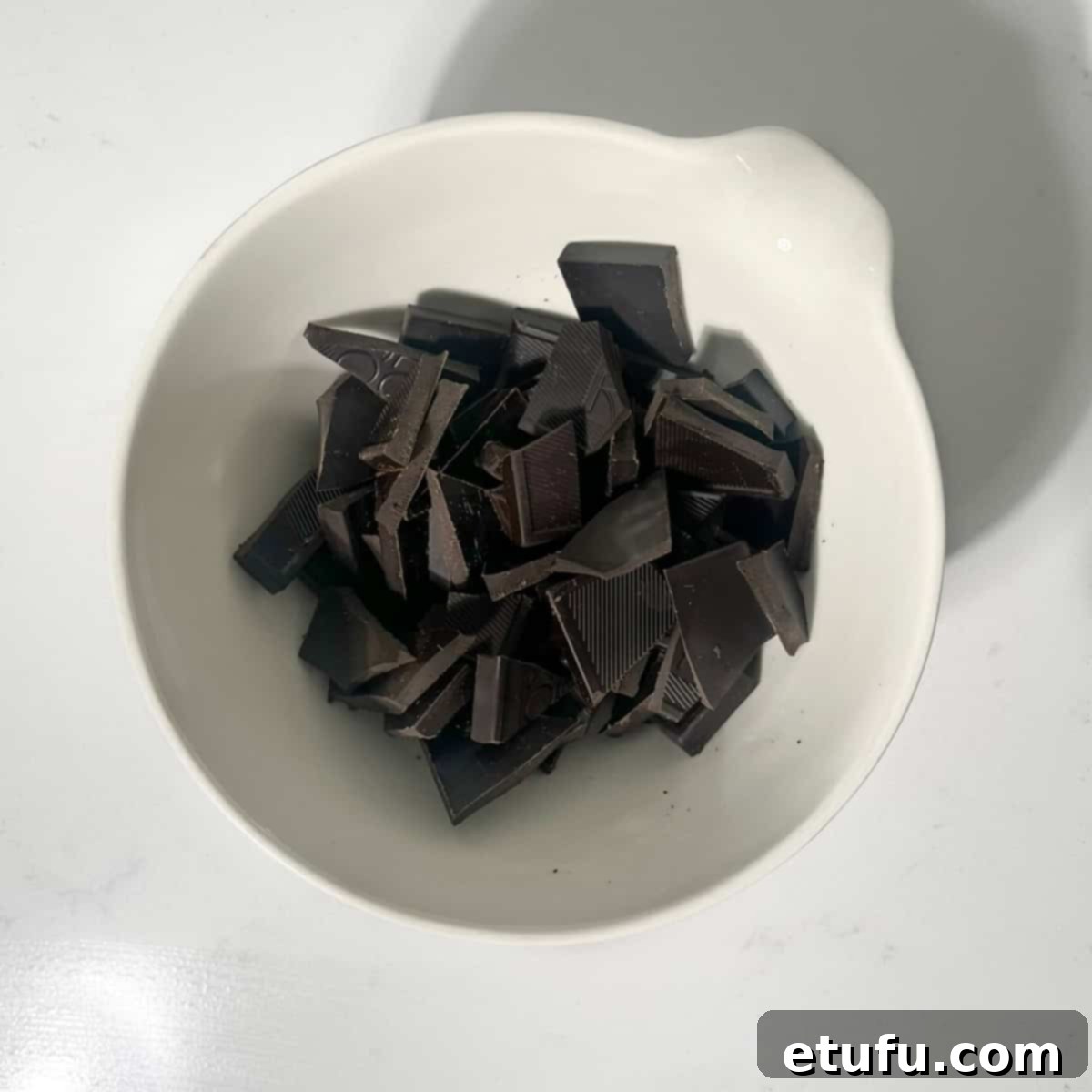 Broken up pieces of dark chocolate in a small white bowl, ready for melting.