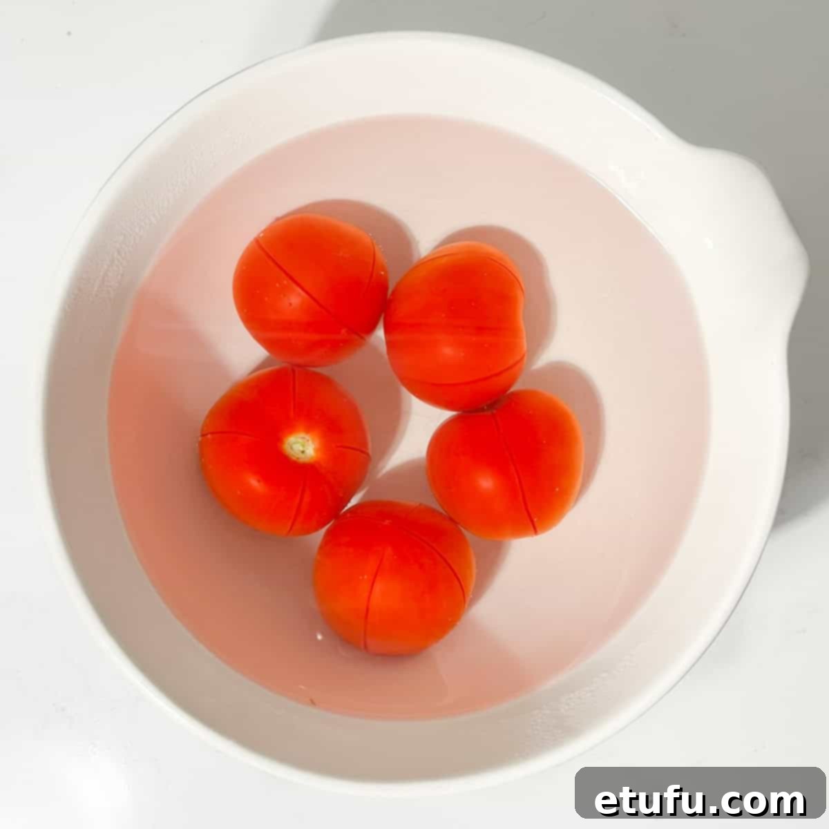 Scored tomatoes soaking in boiled water in a white bowl.