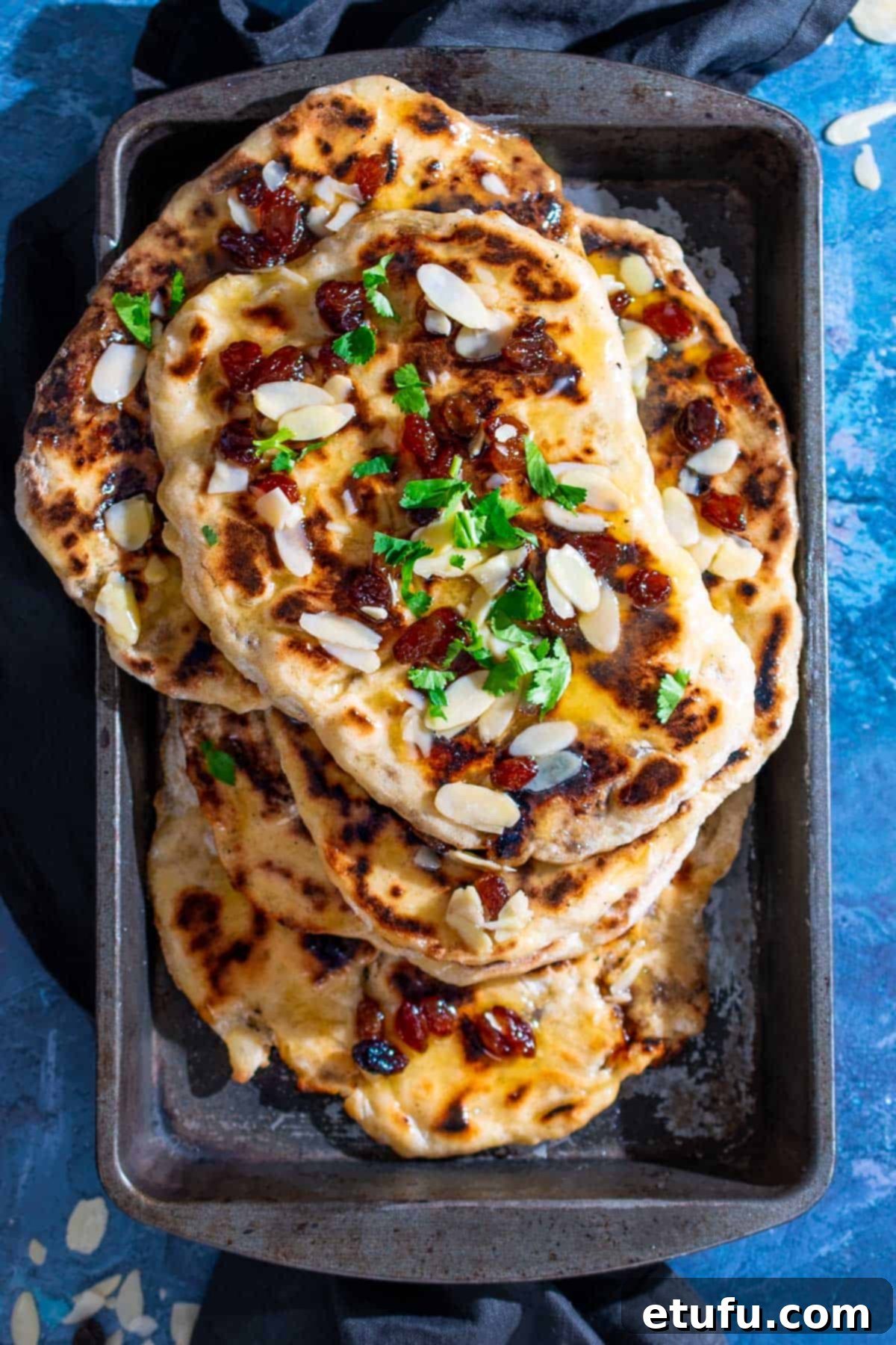 A pile of golden-brown Peshwari naans in a baking dish, ready to be served, on a vibrant blue background.