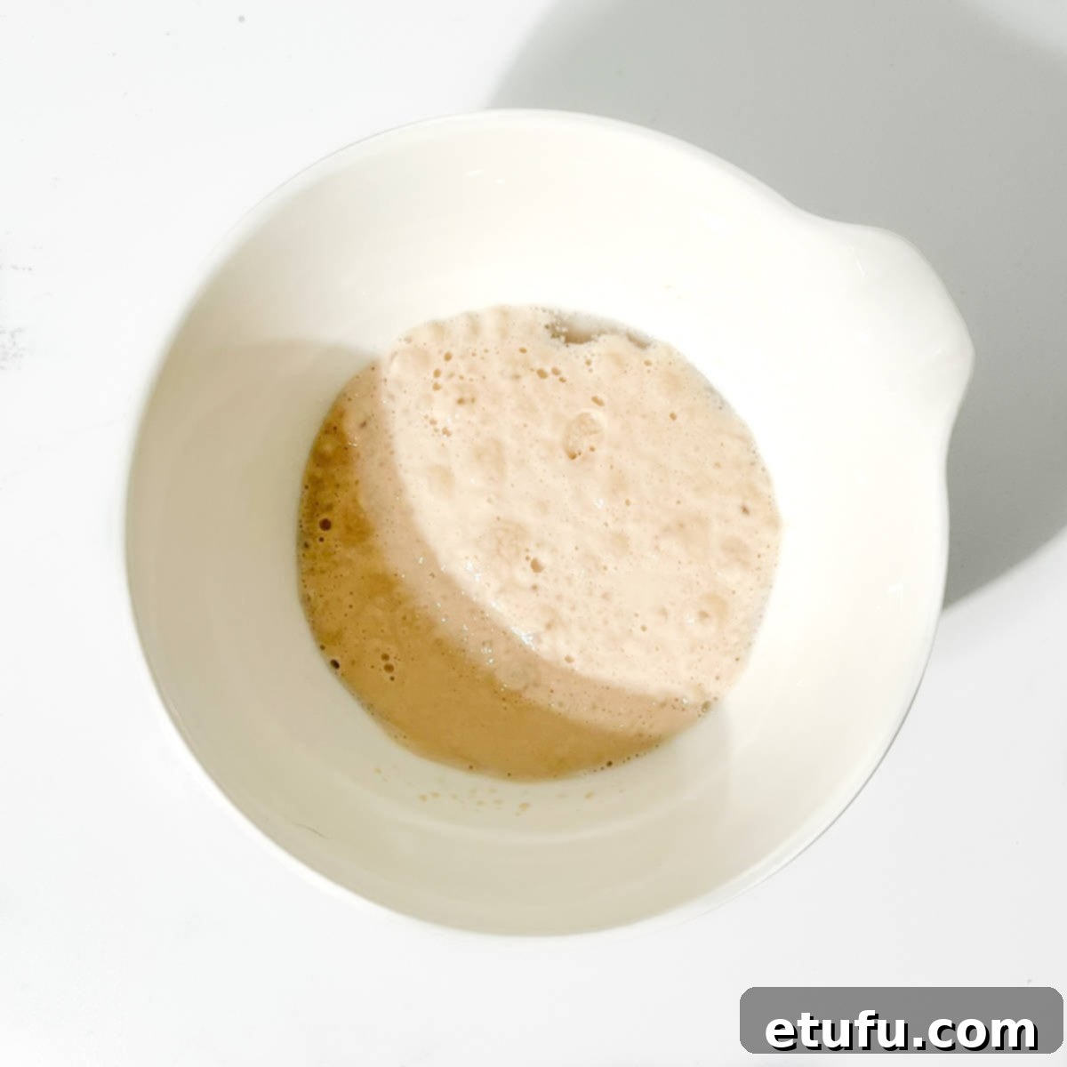 Close-up of activated yeast mixture in a small white bowl, showing a frothy layer on the surface.