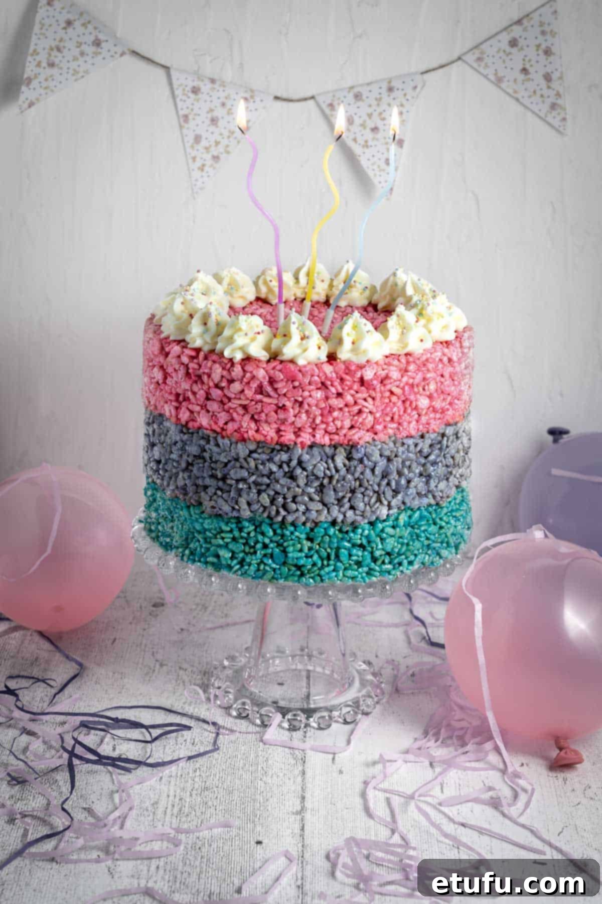 A three layered Rice Krispie cake on a cake stand, surrounded by balloons.
