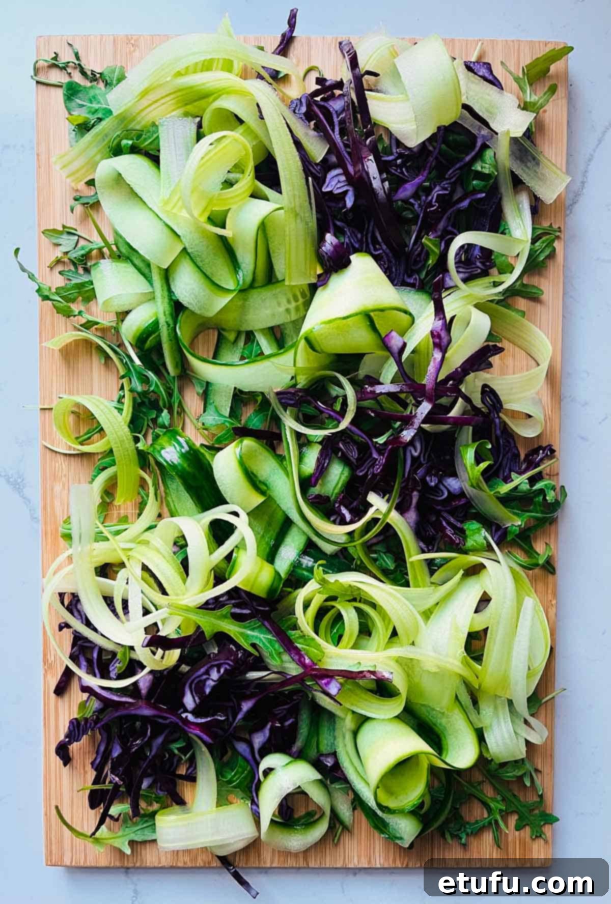 Rocket leaves, red cabbage, and elegant ribbons of celery and cucumber arranged on a wooden board.