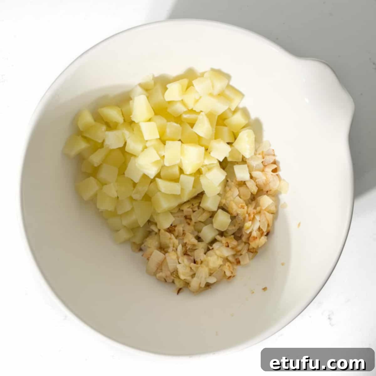 Cooked onions and parboiled potatoes cooling together in a large white mixing bowl, ready for the cheese.