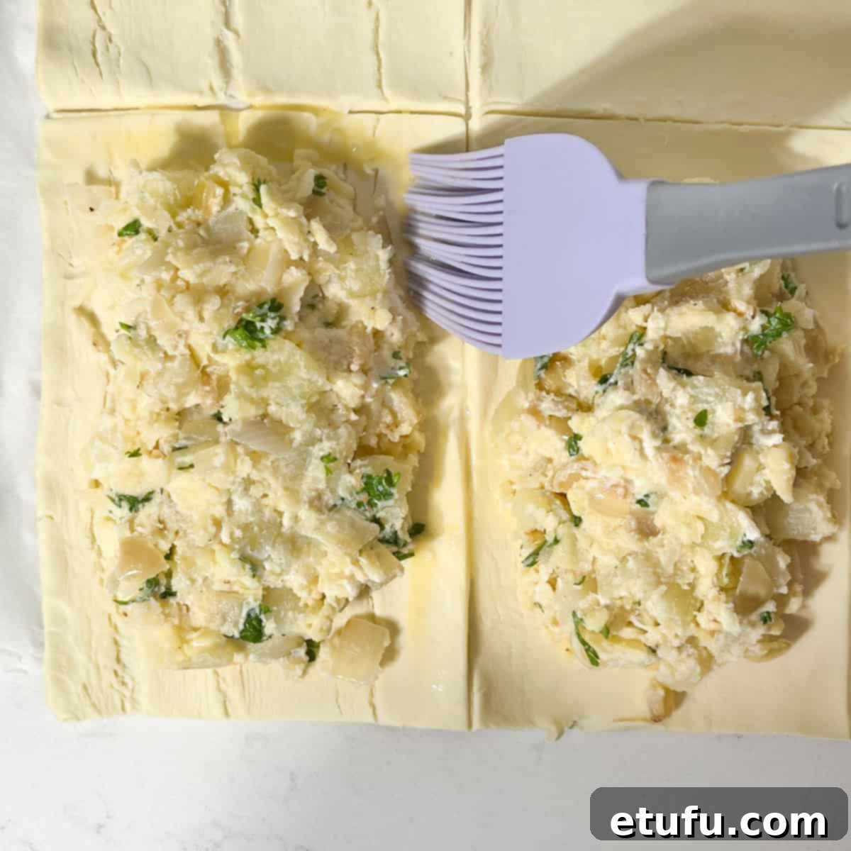 A hand brushing egg wash onto the edges of a puff pastry rectangle with filling, preparing to seal the pasty.