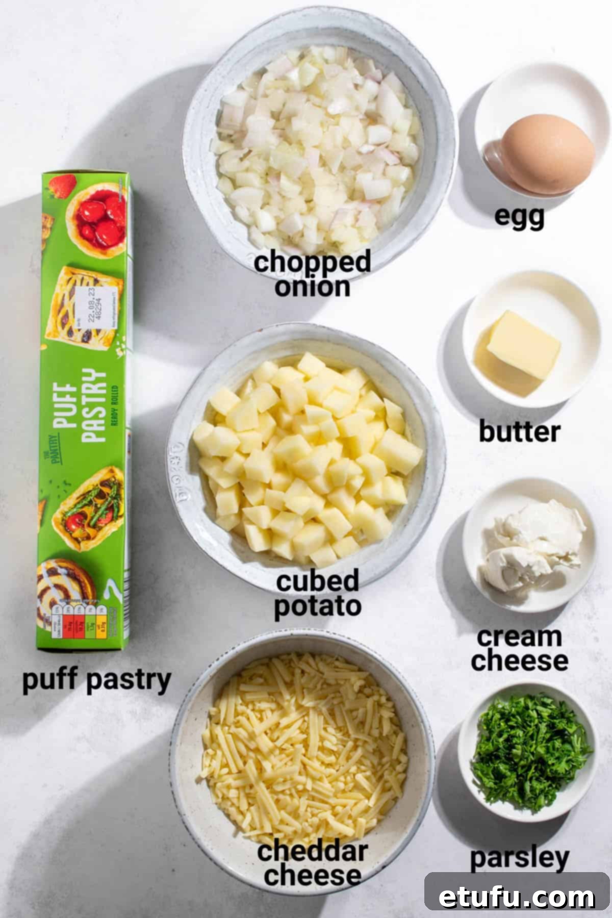 Various ingredients for cheese and onion pasties laid out on a wooden surface, including cheese, onions, potatoes, and a sheet of puff pastry.