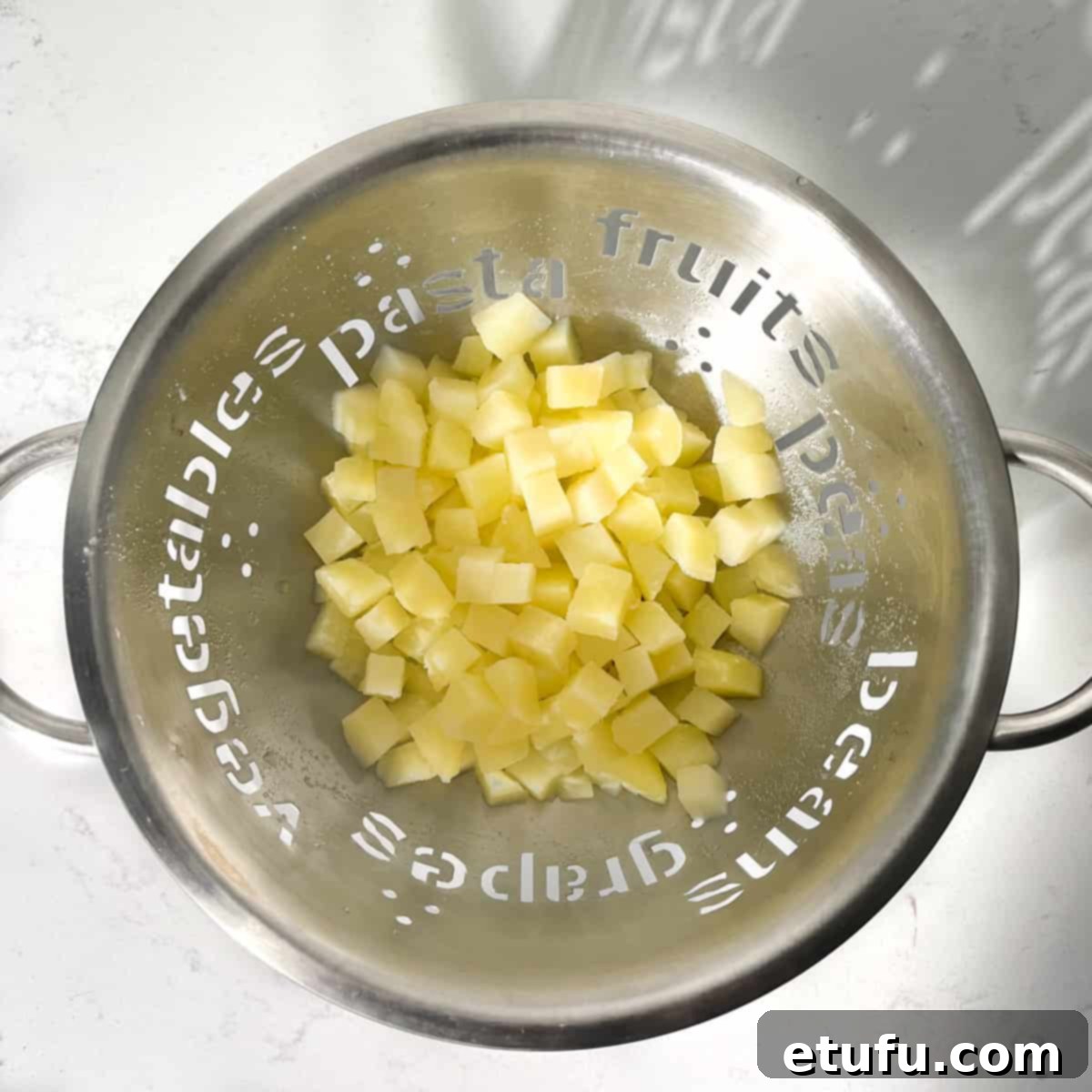 Parboiled potato cubes draining thoroughly in a colander in a kitchen sink.