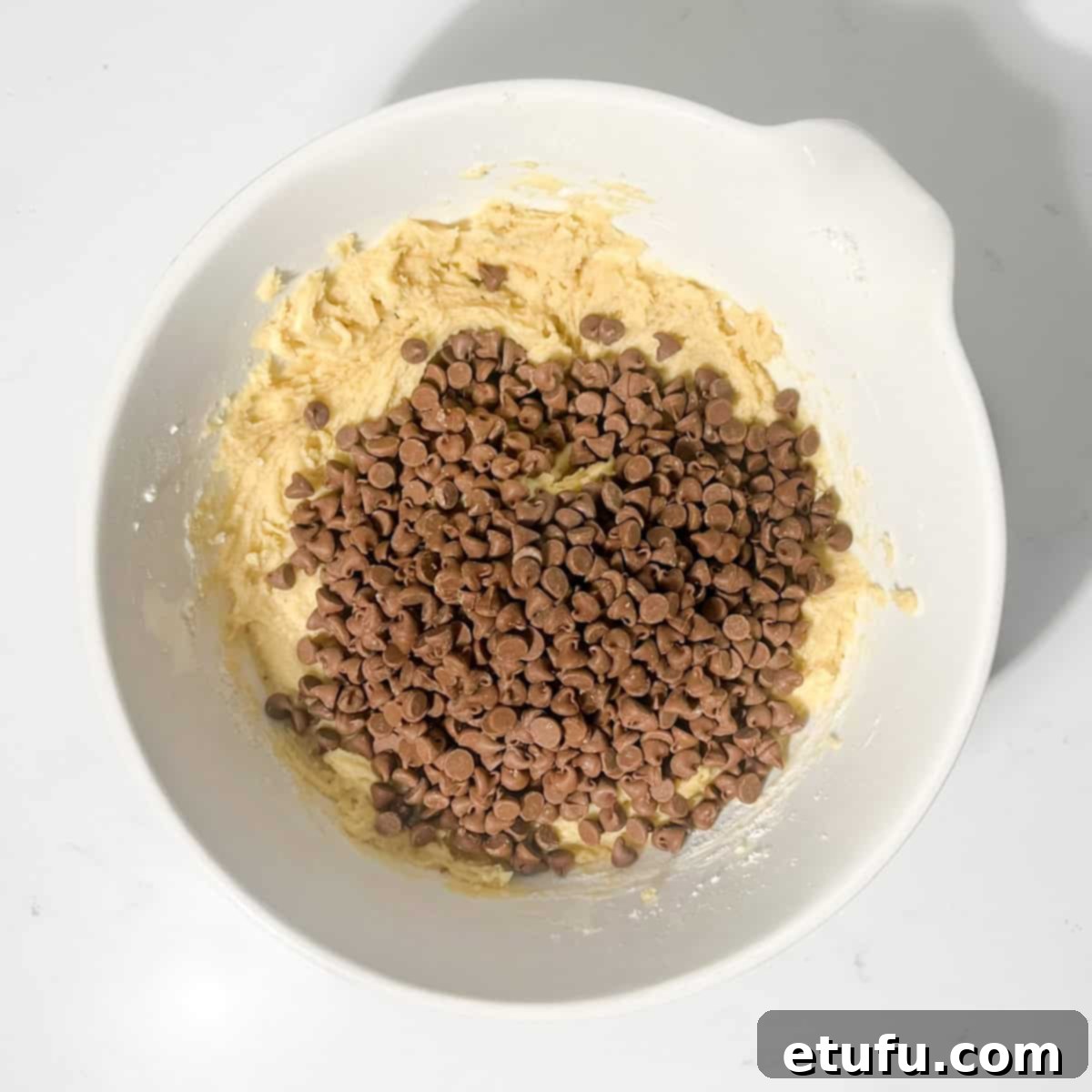 Adding a generous amount of chocolate chips to the prepared cookie dough in a large white bowl.