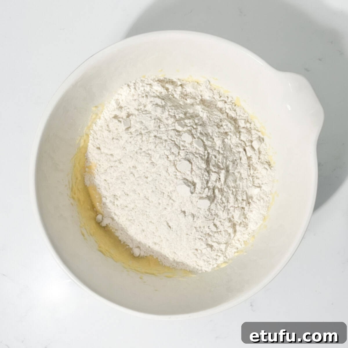 Adding the pre-mixed dry ingredients to the fluffy wet mixture in a large white bowl.