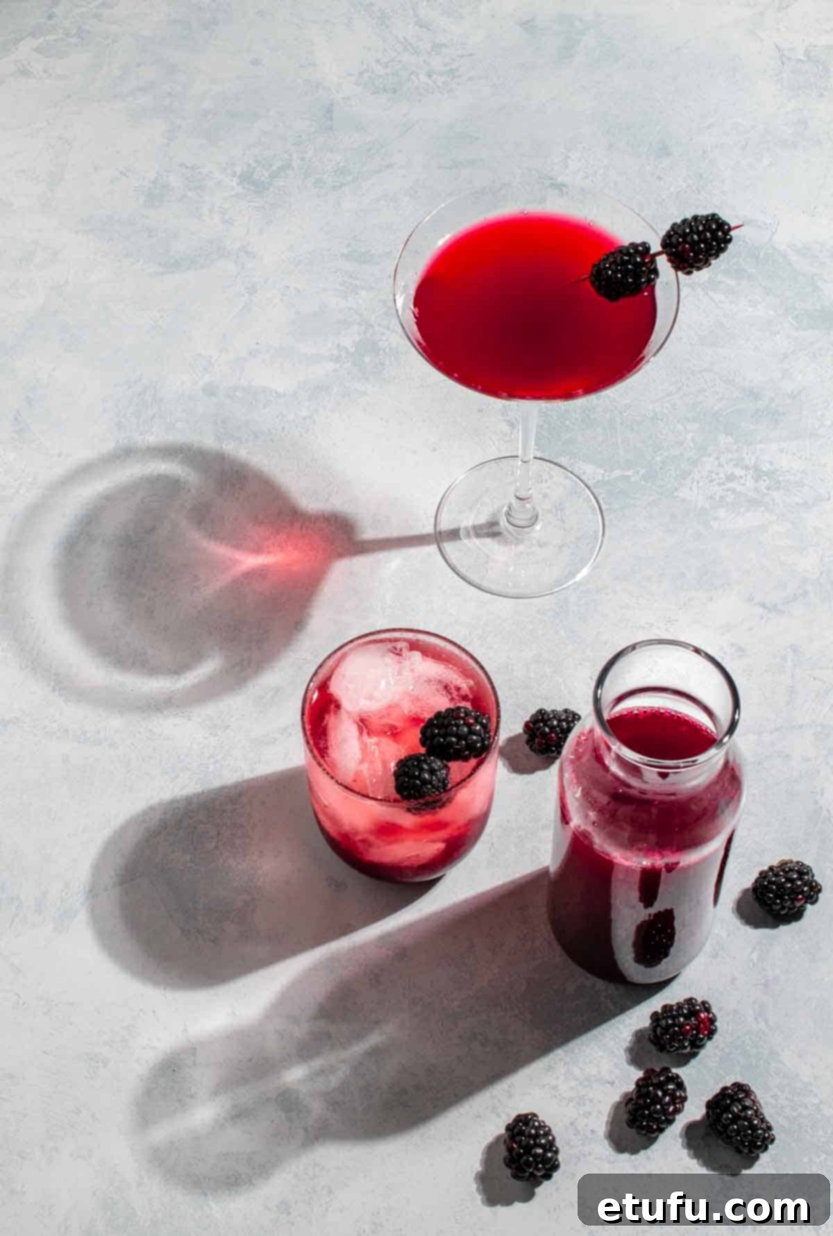 Blackberry simple syrup in a carafe next to a glass of blackberry syrup with lemonade and a blackberry martini.