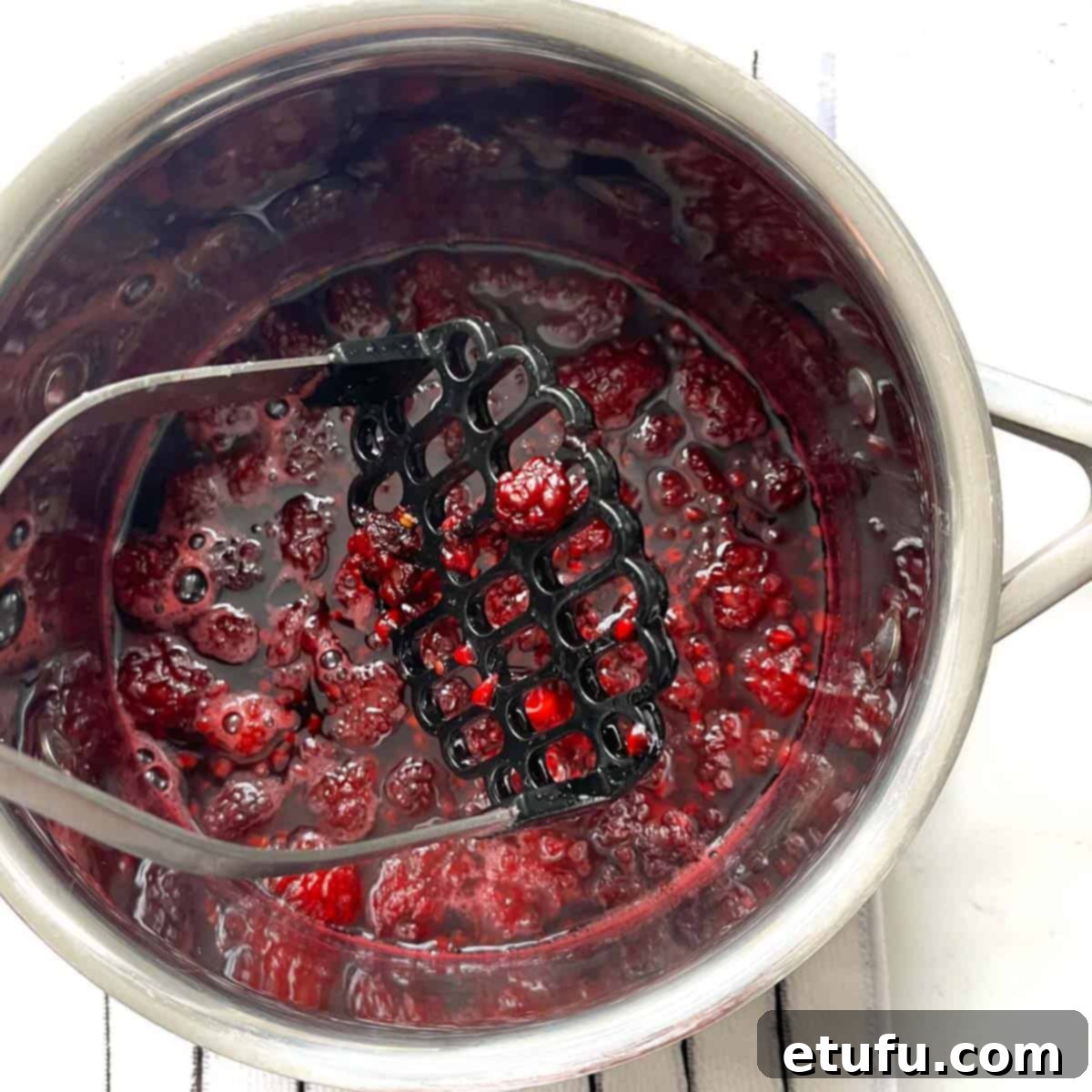 Blackberry simple syrup being mashed with a potato masher in a saucepan.