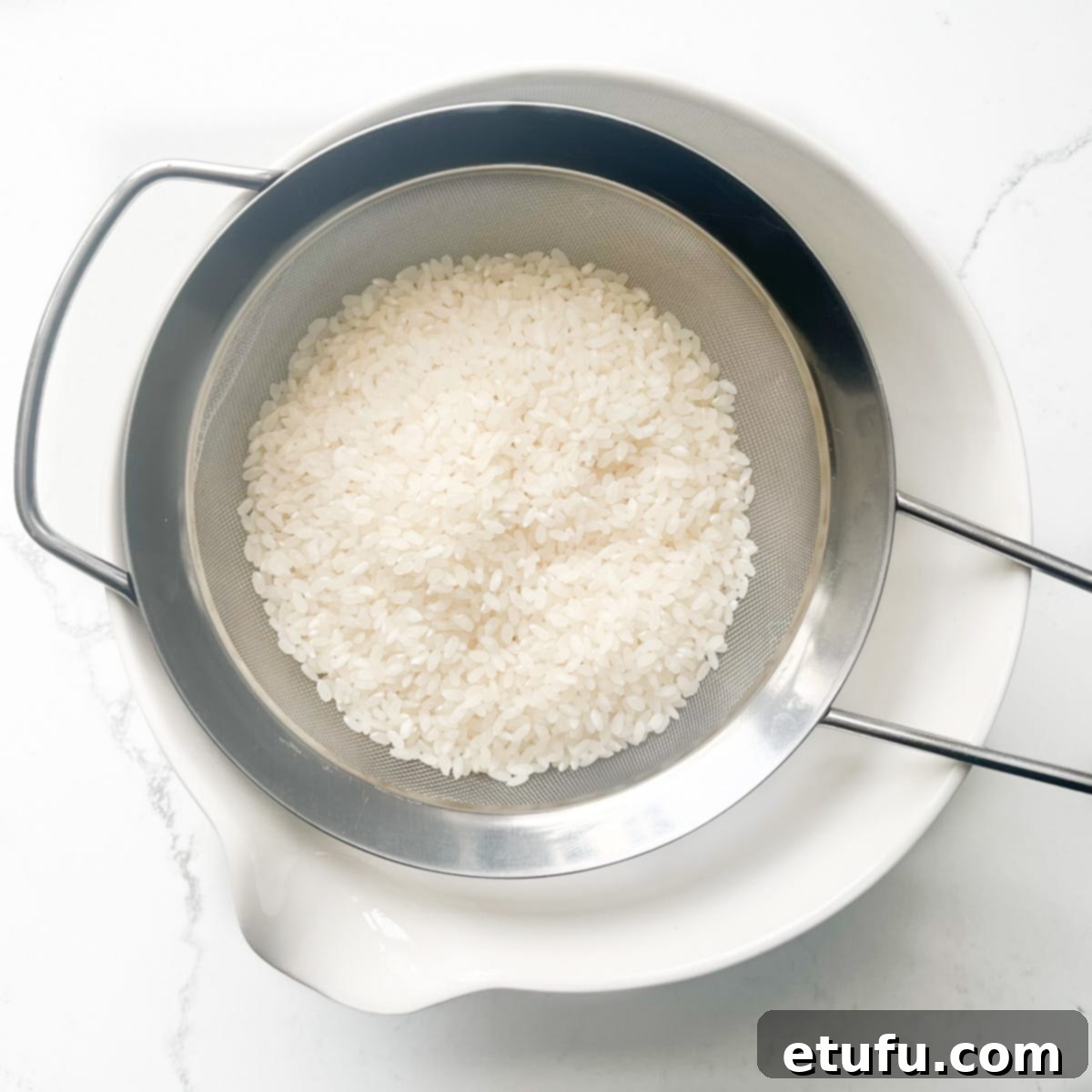 Sushi rice being washed through a sieve over a white bowl. 