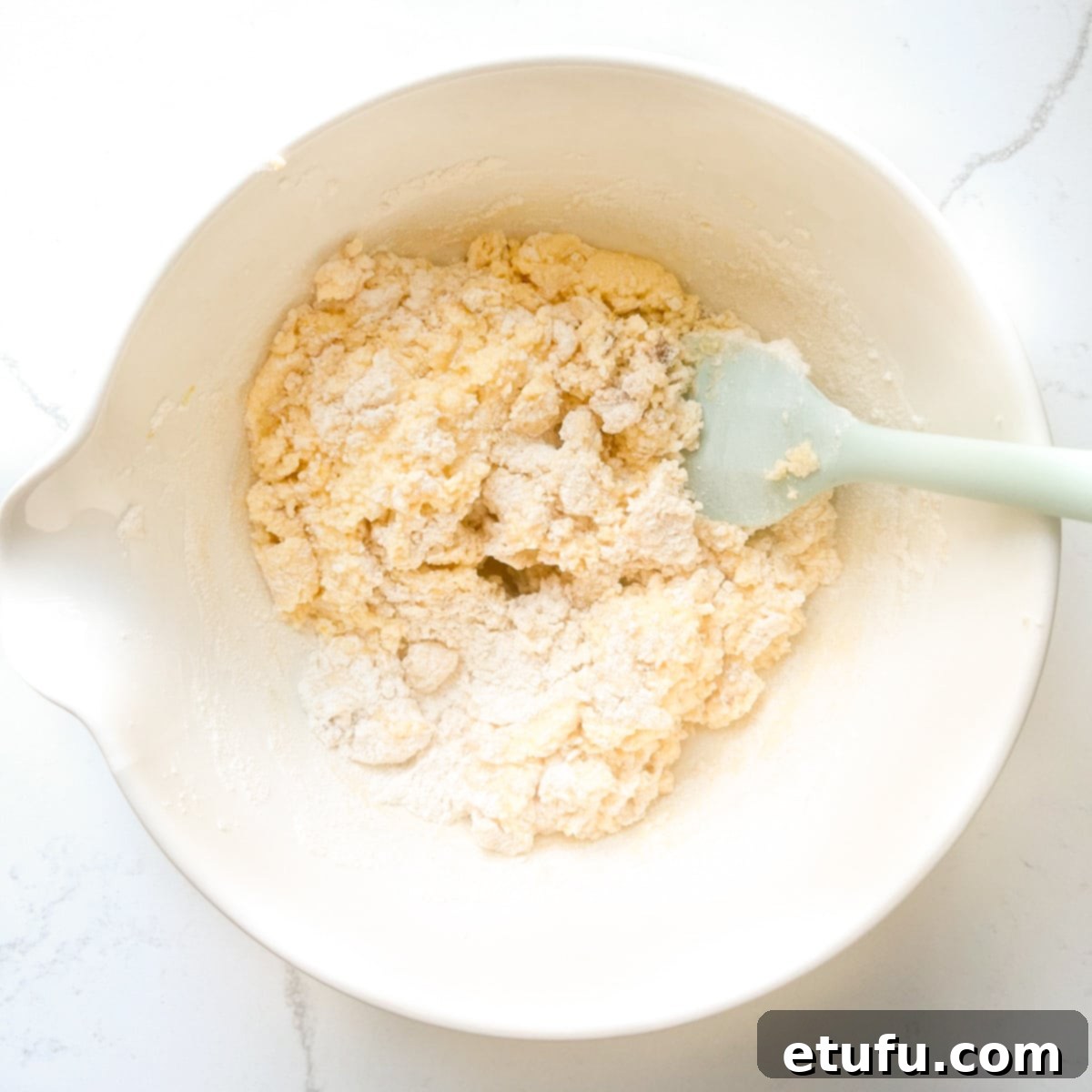 Self-raising flour being gently folded into the cake batter with a spatula in a large mixing bowl.