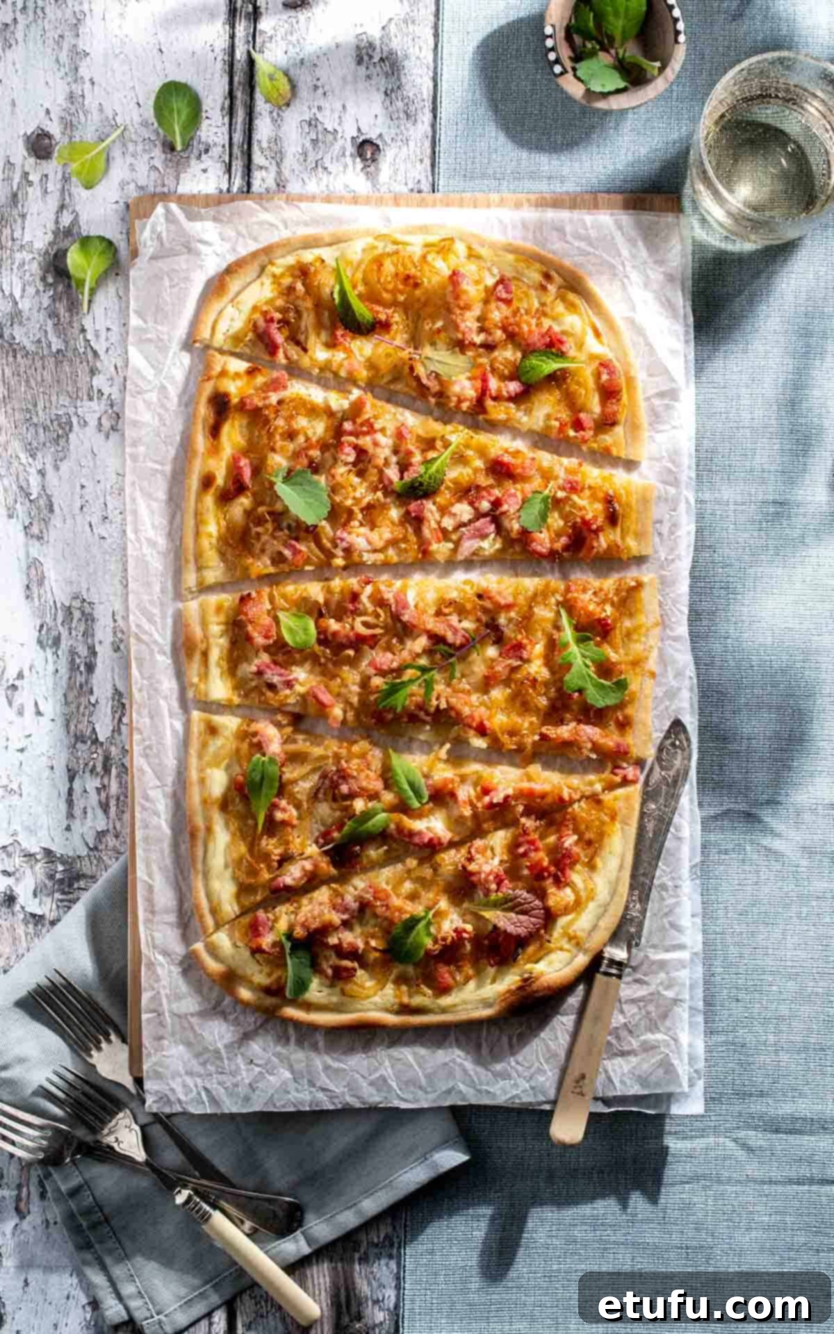 Flammkuchen, a traditional French-German pizza, freshly baked and sliced into wedges, resting on a rustic wooden table. The thin, crispy crust is visible, topped with caramelized onions, bacon lardons, and melted Gruyère cheese, garnished with fresh chives.