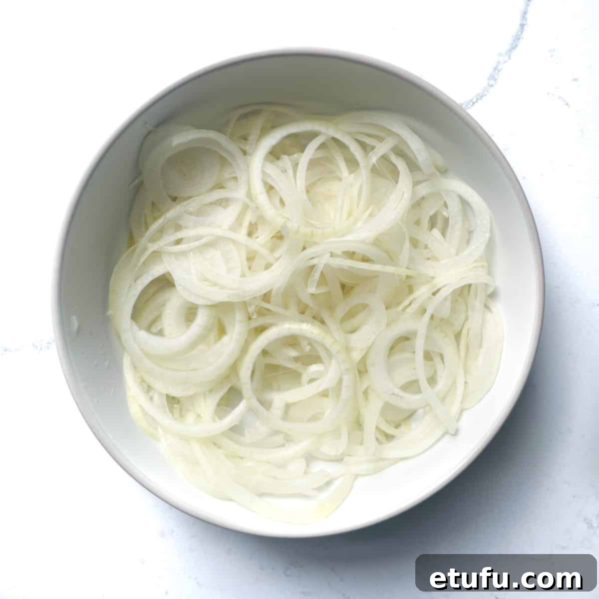 A white bowl filled with finely sliced onions, cut into thin rings, prepared for caramelization and use as a Flammkuchen topping.