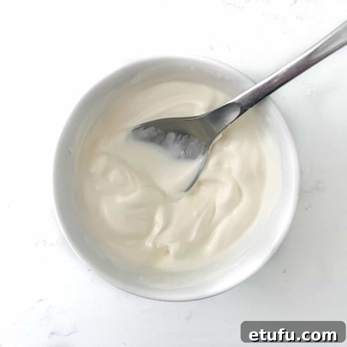 A small white ceramic bowl containing a creamy mixture of sour cream and crème fraîche, expertly combined and ready to be spread on the Flammkuchen base.