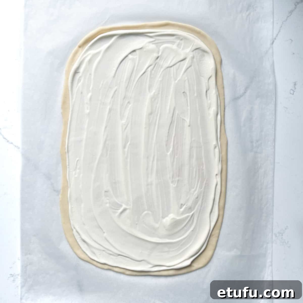 A Flammkuchen pizza base spread evenly with a creamy mixture of sour cream and crème fraîche, leaving a small border, ready for additional toppings.