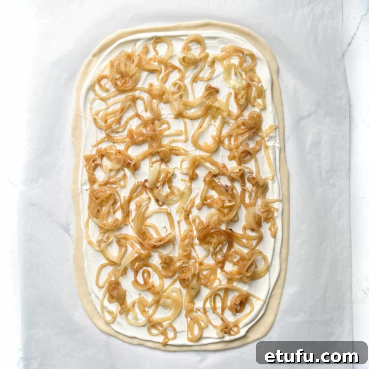 Caramelized onions thinly spread over a cream-covered Flammkuchen pizza base, forming the first layer of toppings.