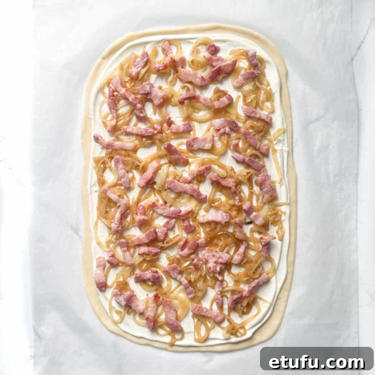 Unbaked Flammkuchen with a generous layer of caramelized onions and scattered bacon lardons on its thin crust, awaiting the cheese layer.