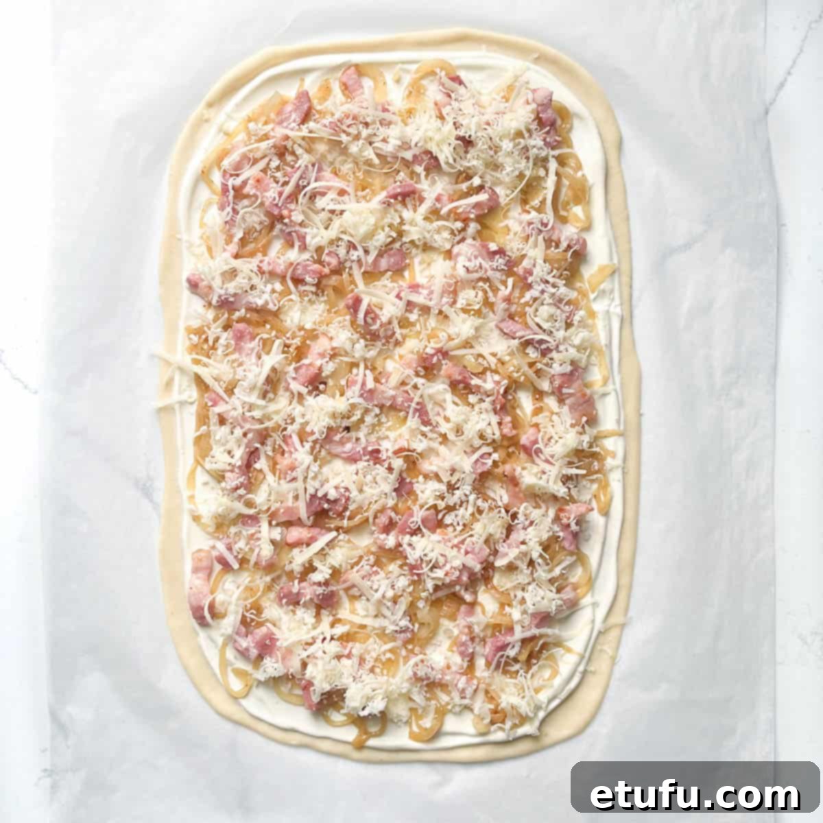 Unbaked Flammkuchen fully topped with caramelized onions, bacon lardons, and a light scattering of grated Gruyère cheese, ready for the oven.