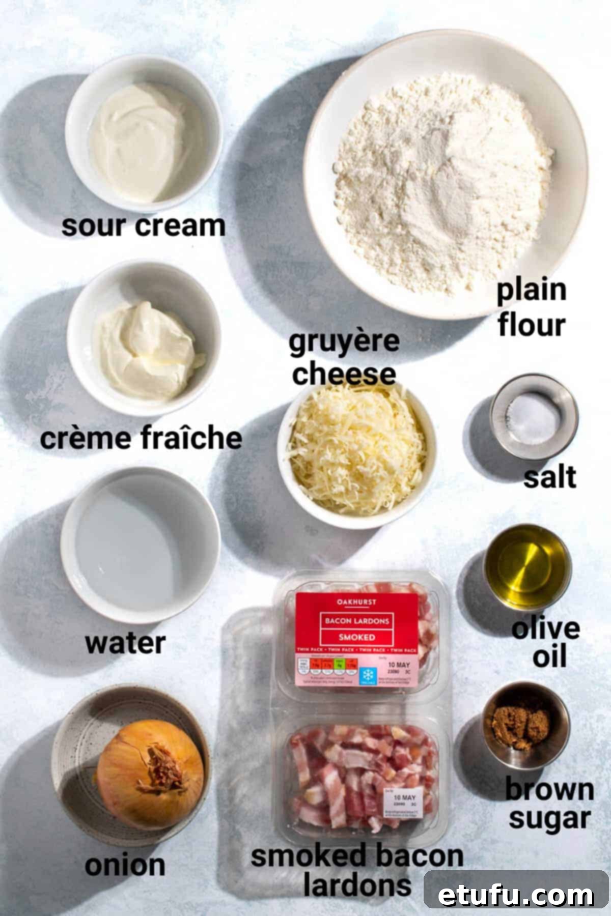 Various Flammkuchen ingredients neatly arranged on a wooden board, including a bowl of sliced onions, smoked bacon lardons, a block of Gruyère cheese, a container of crème fraîche, and a small bowl of fresh chives, ready for preparation.