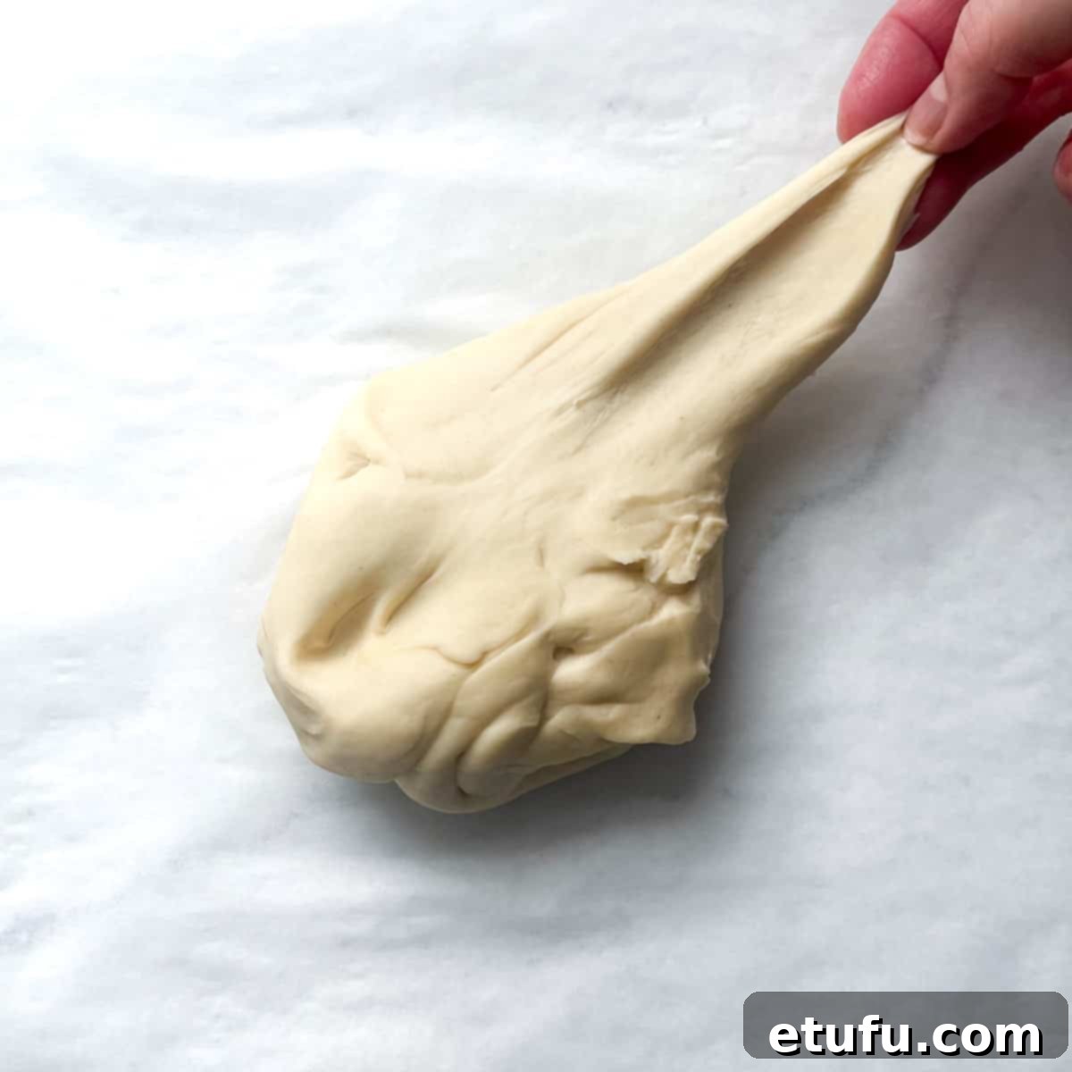 A piece of fully kneaded Flammkuchen dough being stretched to demonstrate its elasticity and smooth texture, indicating it's ready for rolling.