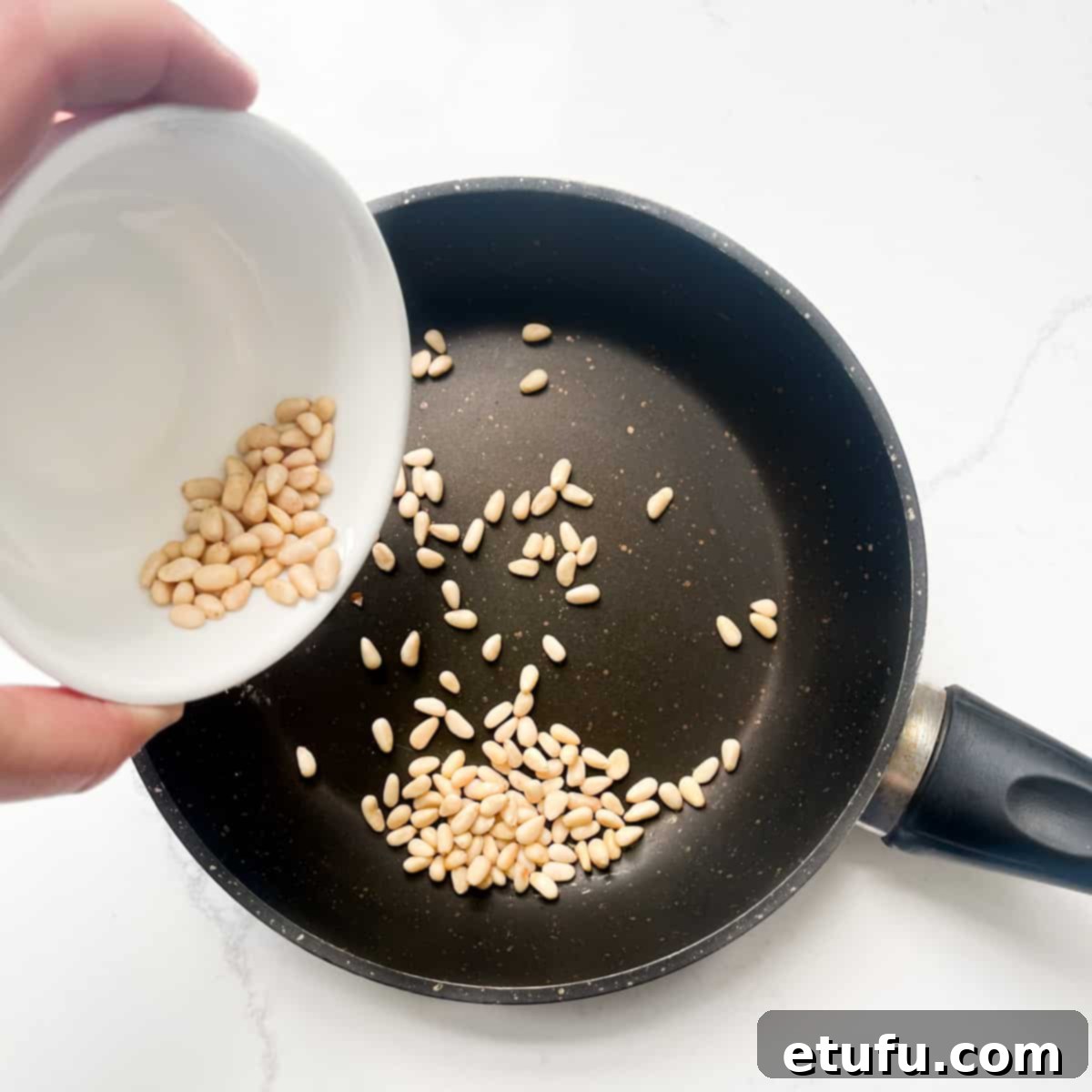 Adding pine nuts to a small frying pan to be roasted until golden brown.