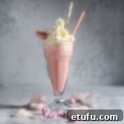 A large glass of strawberry milkshake with whipped cream in a milkshake glass surrounded by cotton candy.
