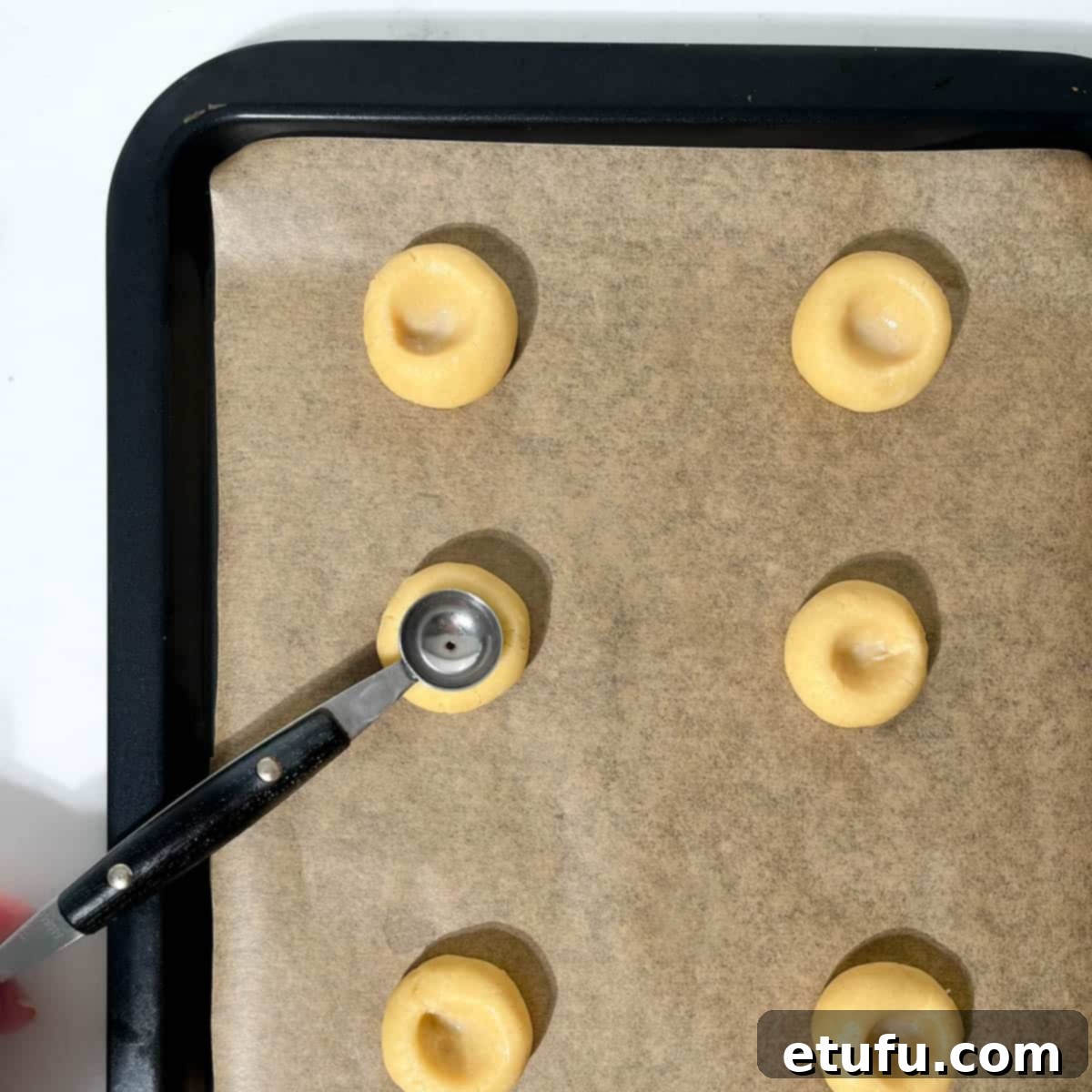 Indenting round dough balls for the thumbprint icing cookies with a small melon baller on a baking tray.