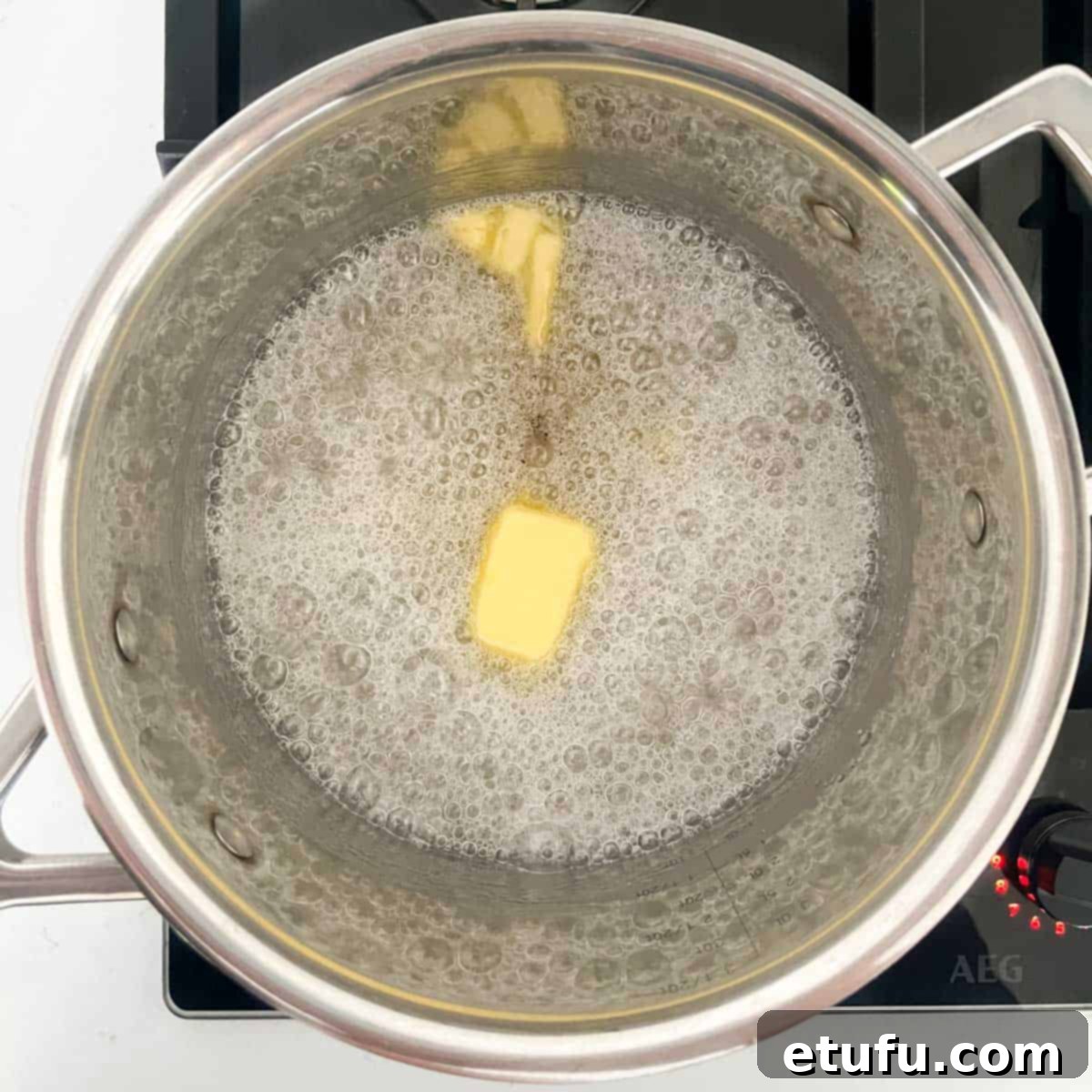 Unsalted butter and vanilla extract being added to the simmering sugar and water mixture in a saucepan, forming the base for homemade peanut brittle.
