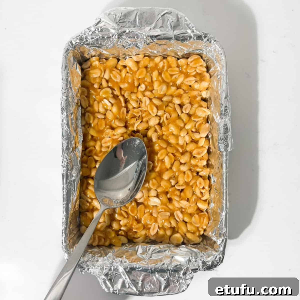 The hot peanut brittle mixture being smoothed evenly into a foil-lined baking tray using the back of a metal spoon.