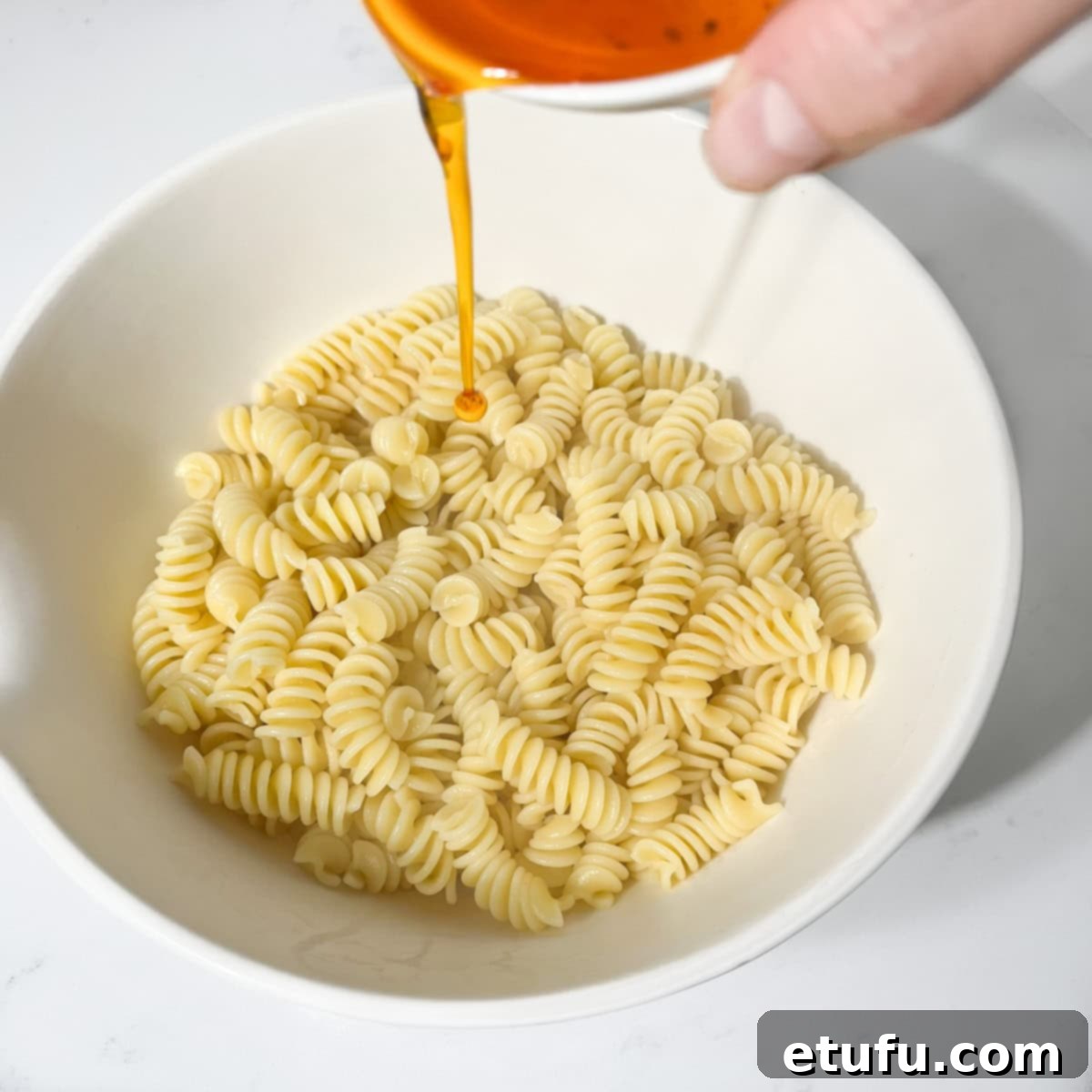 Adding some of the reserved red pepper oil to the cooked pasta in a large white bowl.