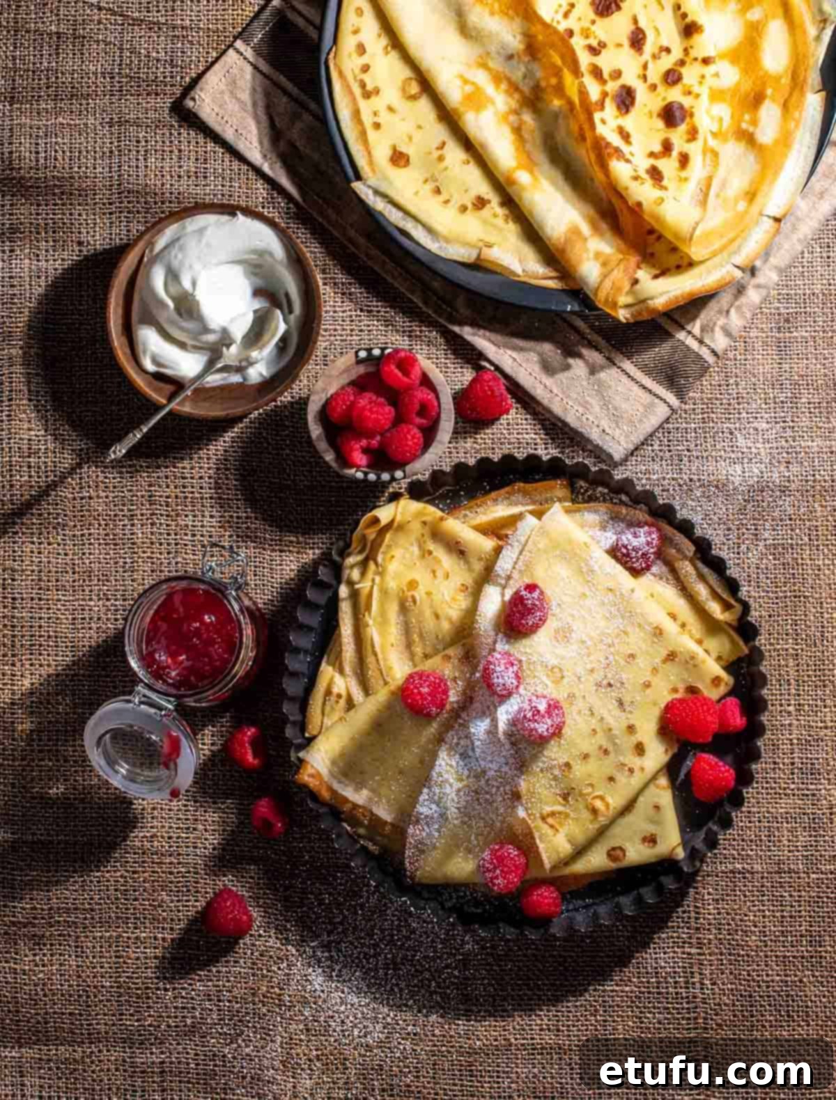 Pandekager folded in triangles in a black, round backing dish, served with raspberries, icing sugar, jam and cream.