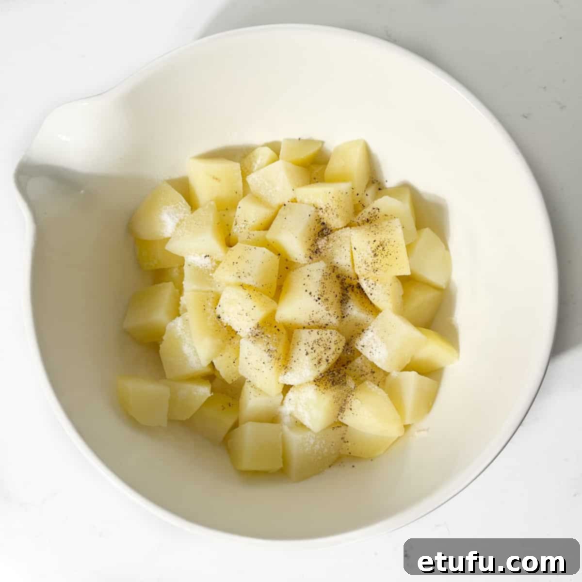 Potato cubes in a large white bowl with olive oil, salt and pepper.