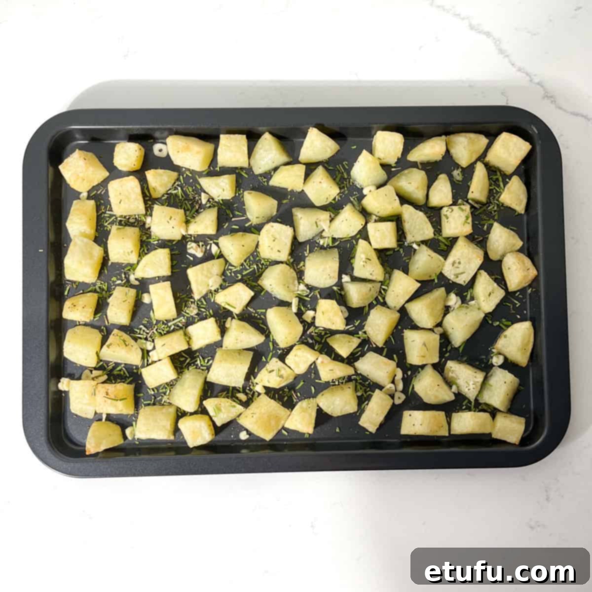 Partially cooked parmentier potatoes on a baking tray, sprinkled with garlic and rosemary.